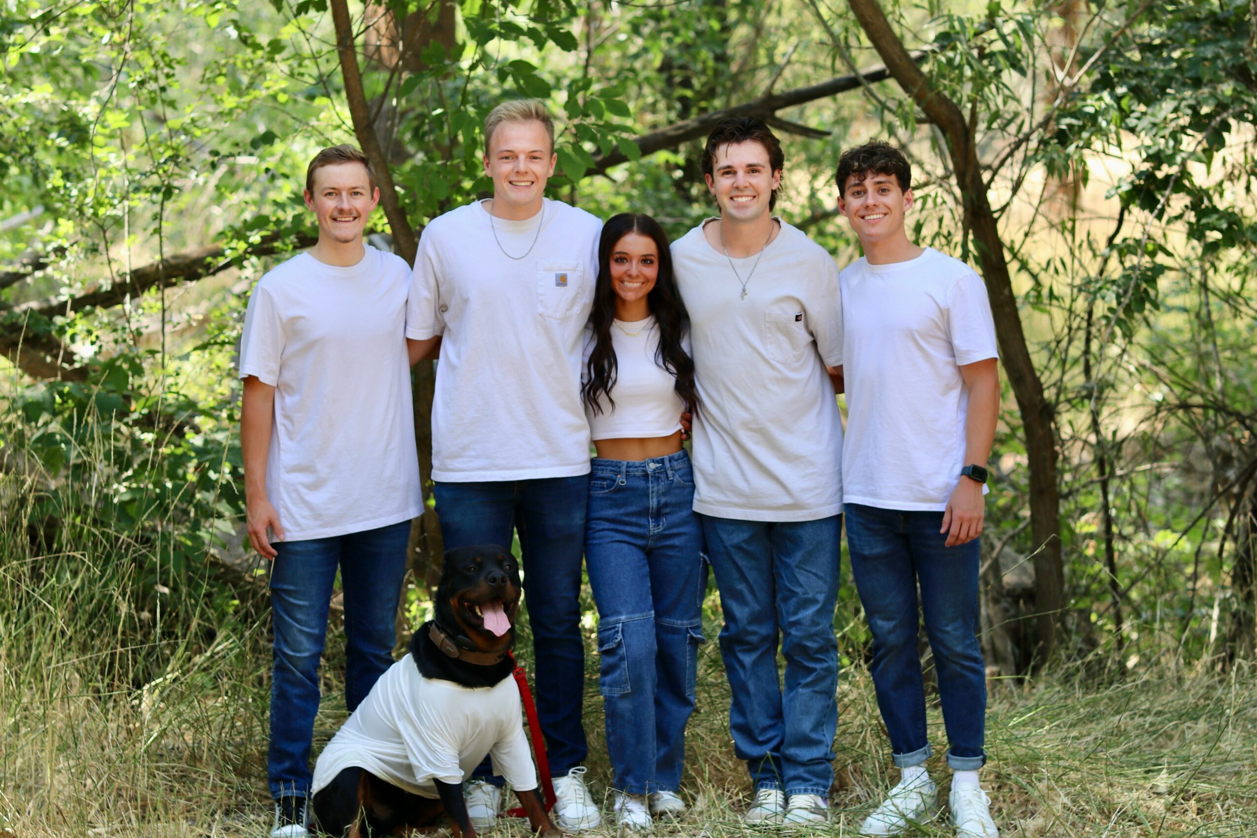 Group of five people standing outdoors in a wooded area, smiling, with a dog in front wearing a white shirt.