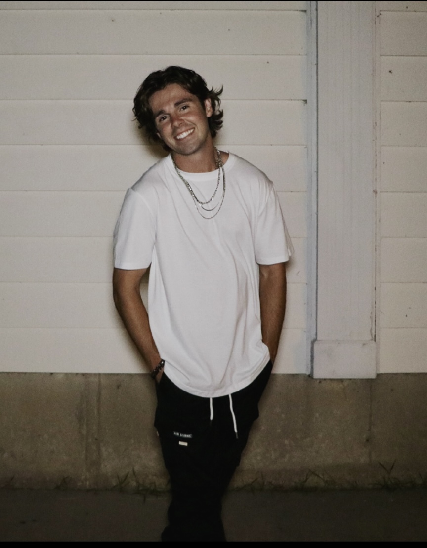 Young man with curly hair smiling, wearing a white t-shirt and layered necklaces, standing against a white wall.