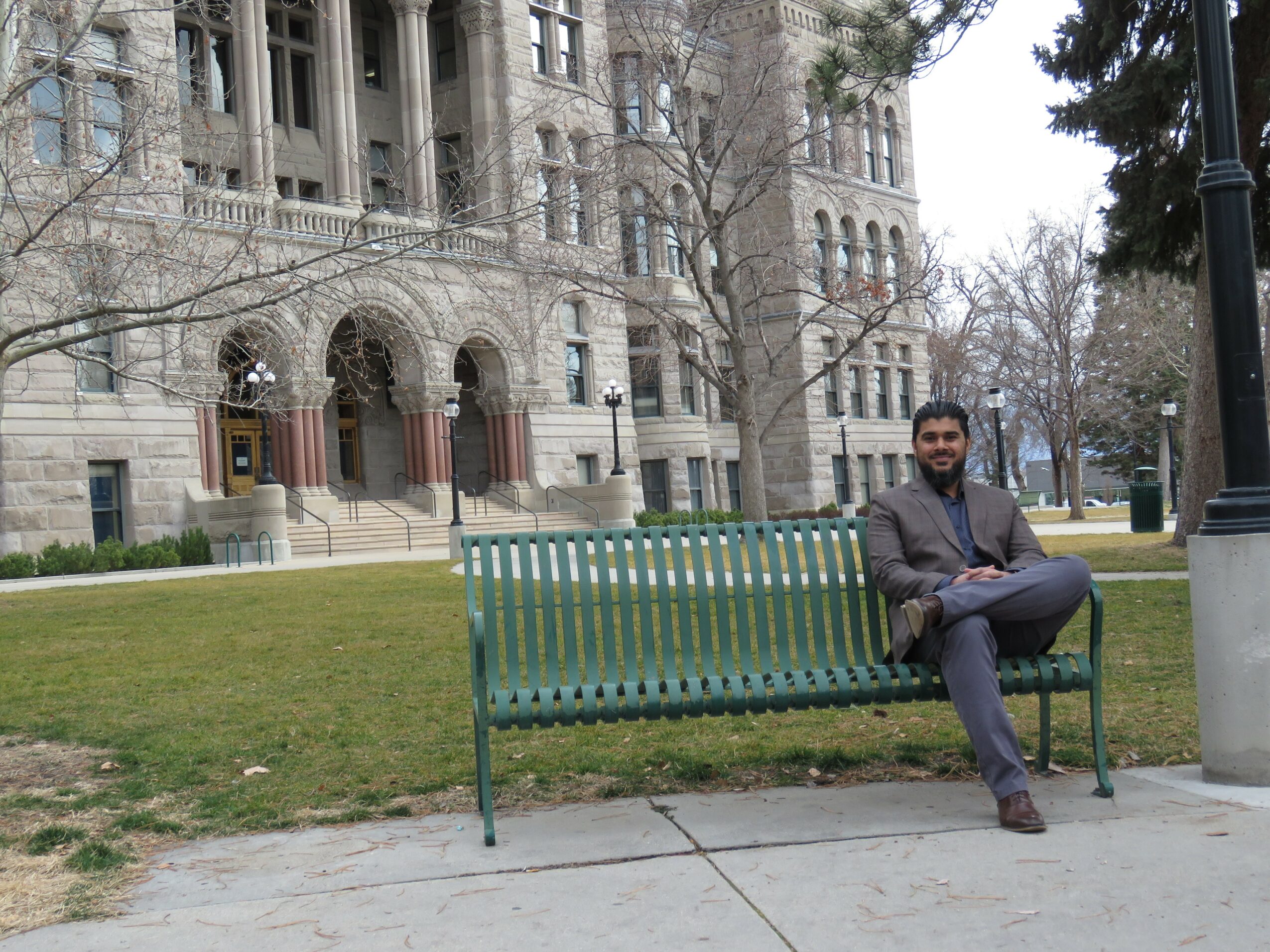 Man sitting on a green park bench in front of a historic stone building with arched windows and trees.