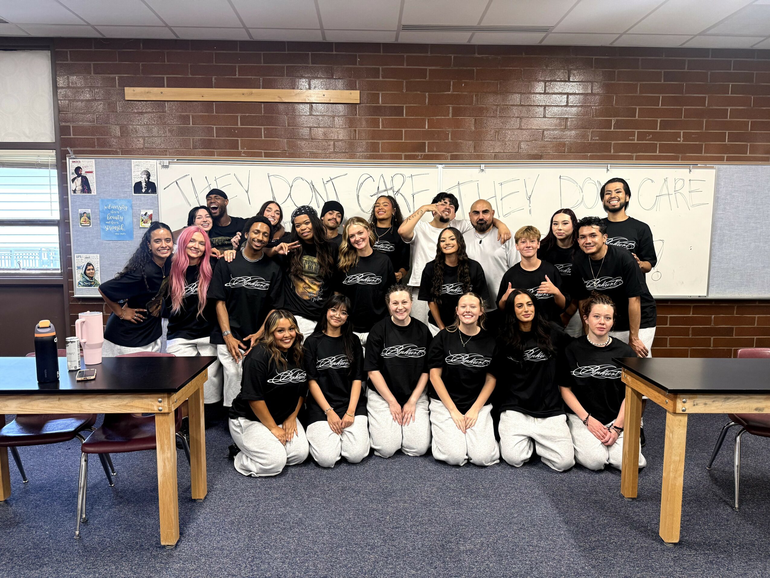 Group of students in black shirts and white pants posing in classroom with whiteboard and brick wall.