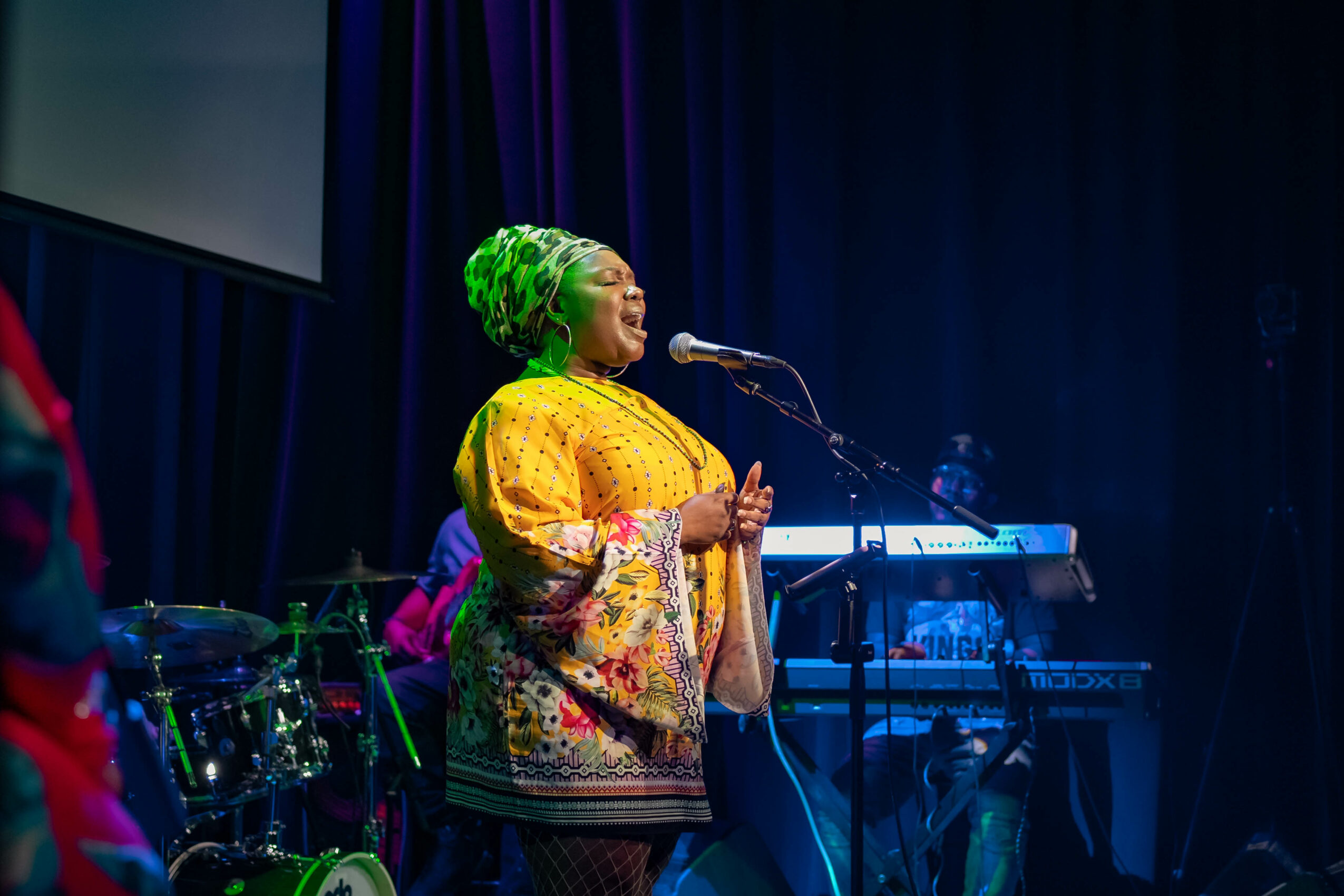Woman with green hair singing into a microphone on stage with musical instruments in background.
