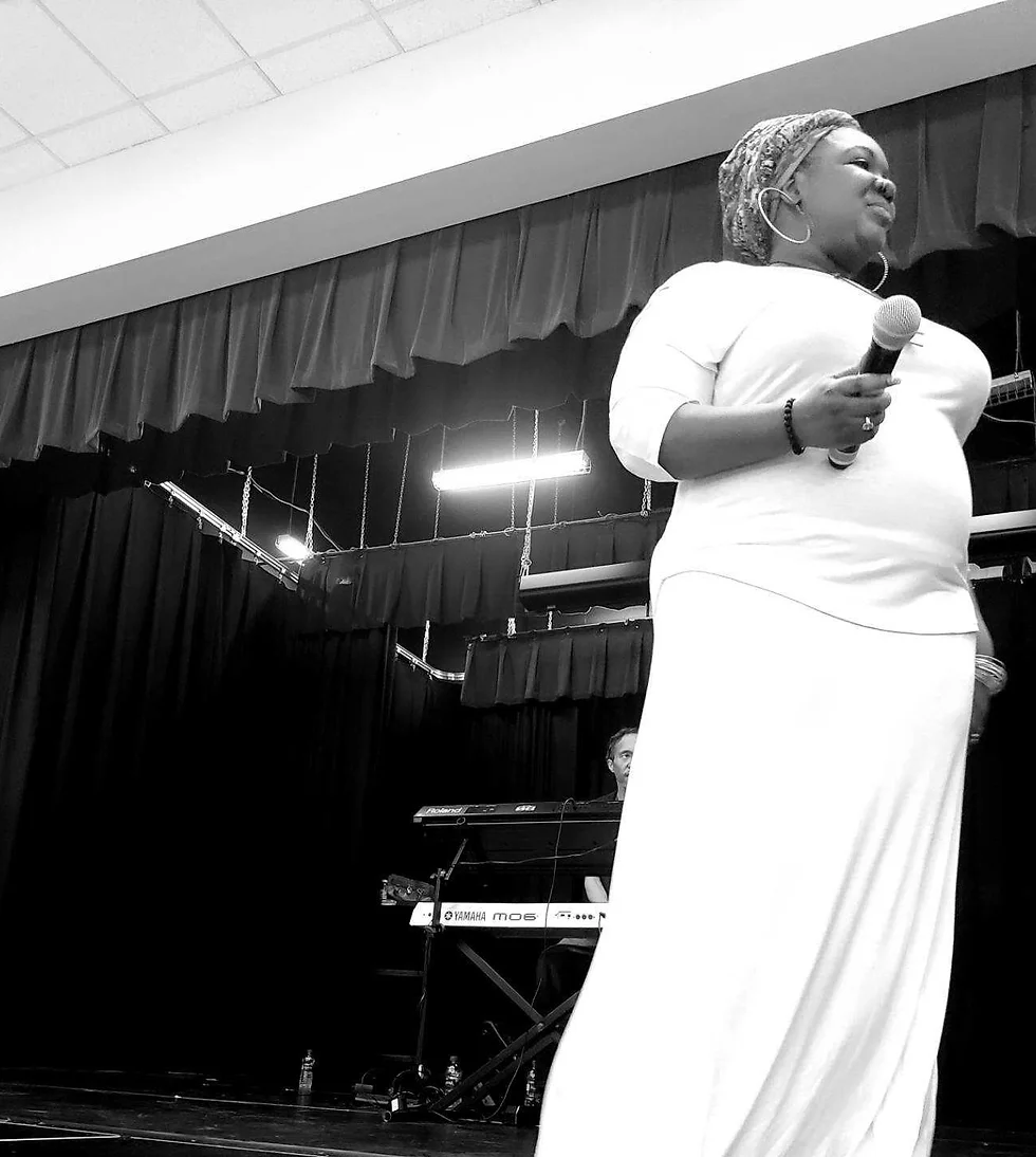 Woman holding a microphone on stage, with a keyboard and a person in the background, in a black and white photo.