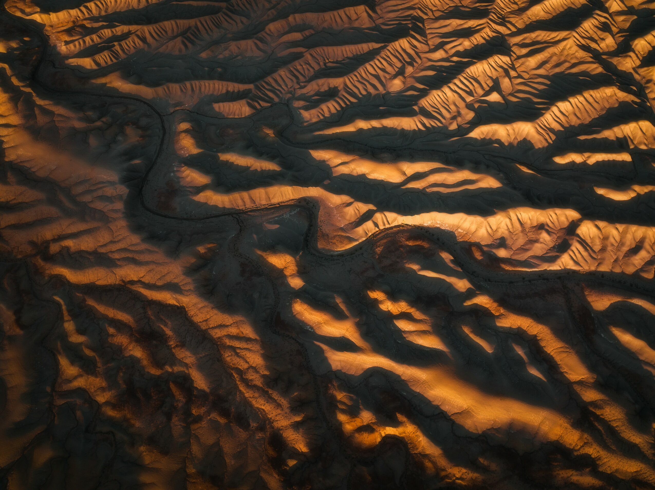 Aerial view of rugged canyon terrain with deep shadows and winding river channels, illuminated by warm sunlight.