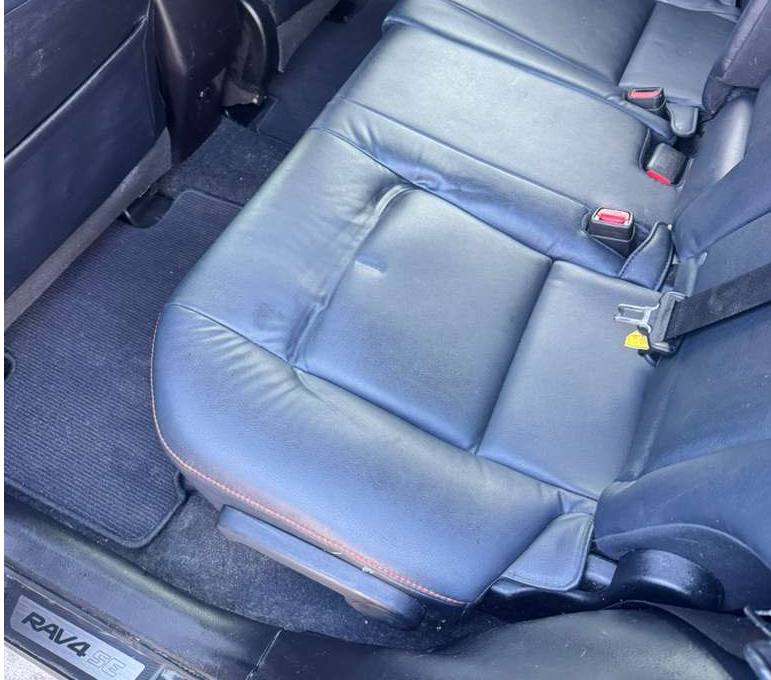 Car back seat with black leather upholstery and seat belts, floor mat visible, part of door frame in view.