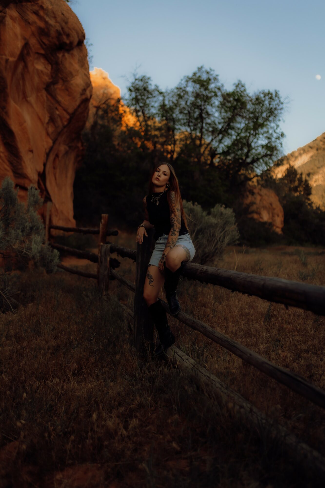 Person sitting on a wooden fence outdoors with trees and rocks, sunset lighting, and a clear sky in the background.