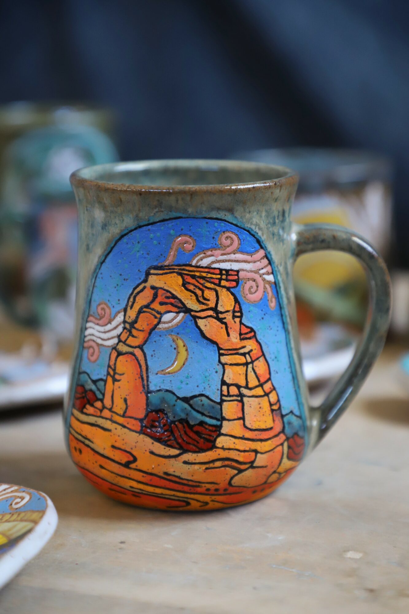 Ceramic mug with desert landscape and arch design, featuring a blue sky, moon, and rocky formations.