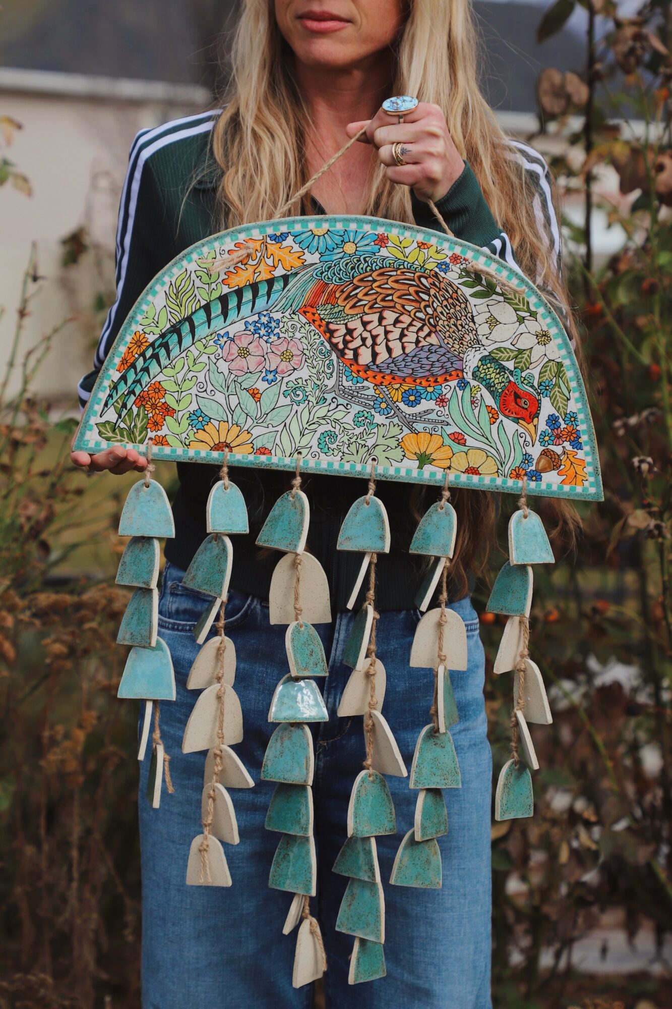 Person holding a colorful, semi-circular fabric bag with tassels, outdoors with plants in the background.