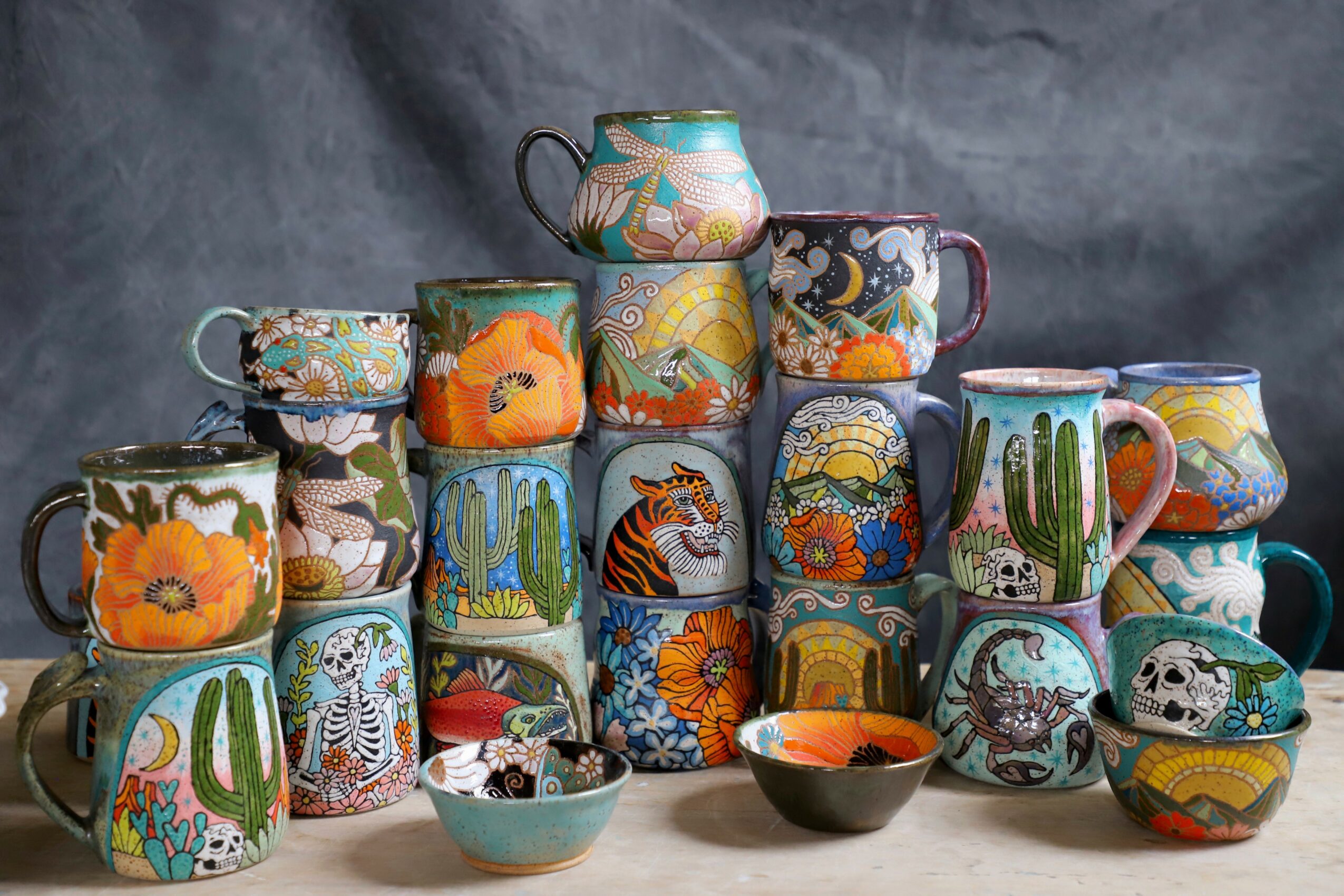 Stacked colorful mugs and bowls with various painted designs, including animals, plants, and abstract patterns, on a gray background.