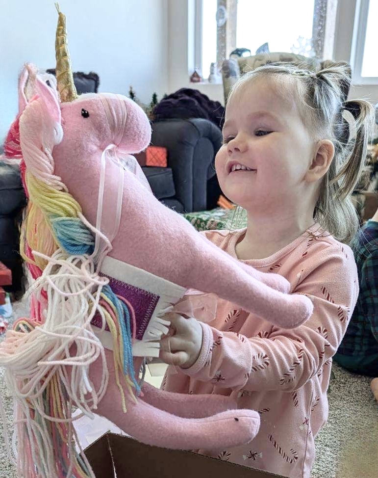 Young girl smiling and playing with a pink stuffed unicorn in a room with large windows and various objects