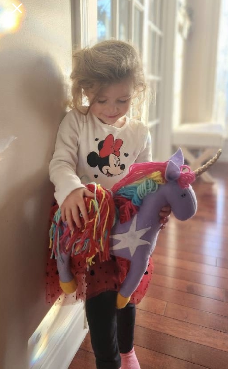 Child with curly hair playing with a colorful unicorn plush toy indoors, sunlight streaming through windows, wooden floor visible.