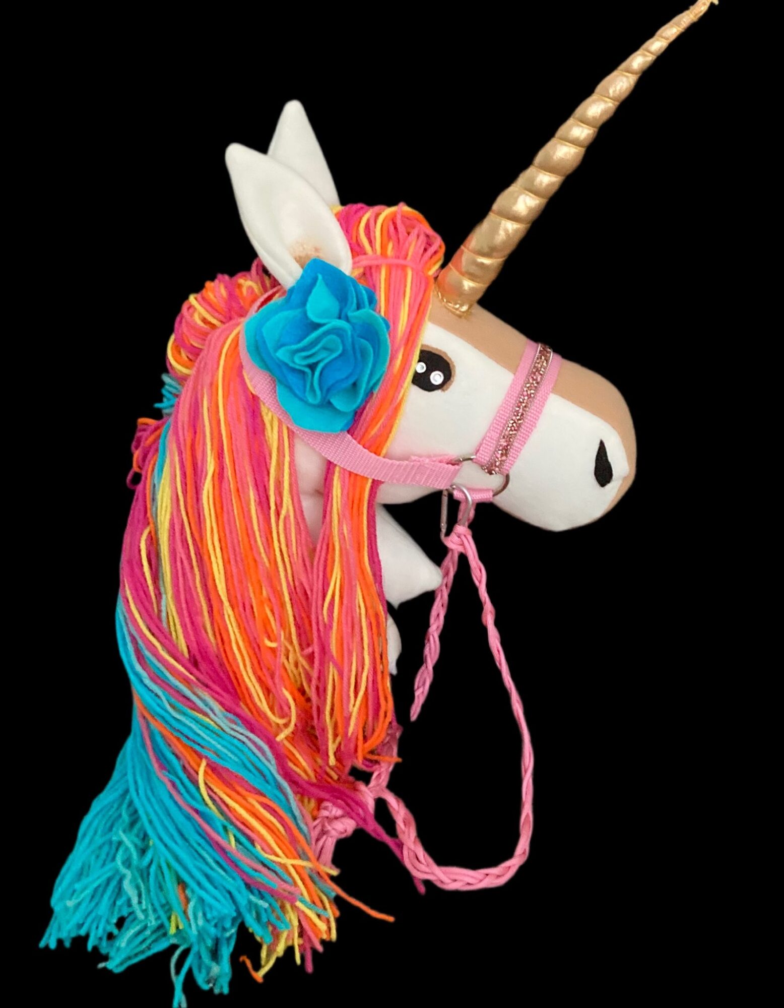 Colorful unicorn plush toy with a golden horn, pink bridle, and rainbow yarn mane and tail, against a black background.