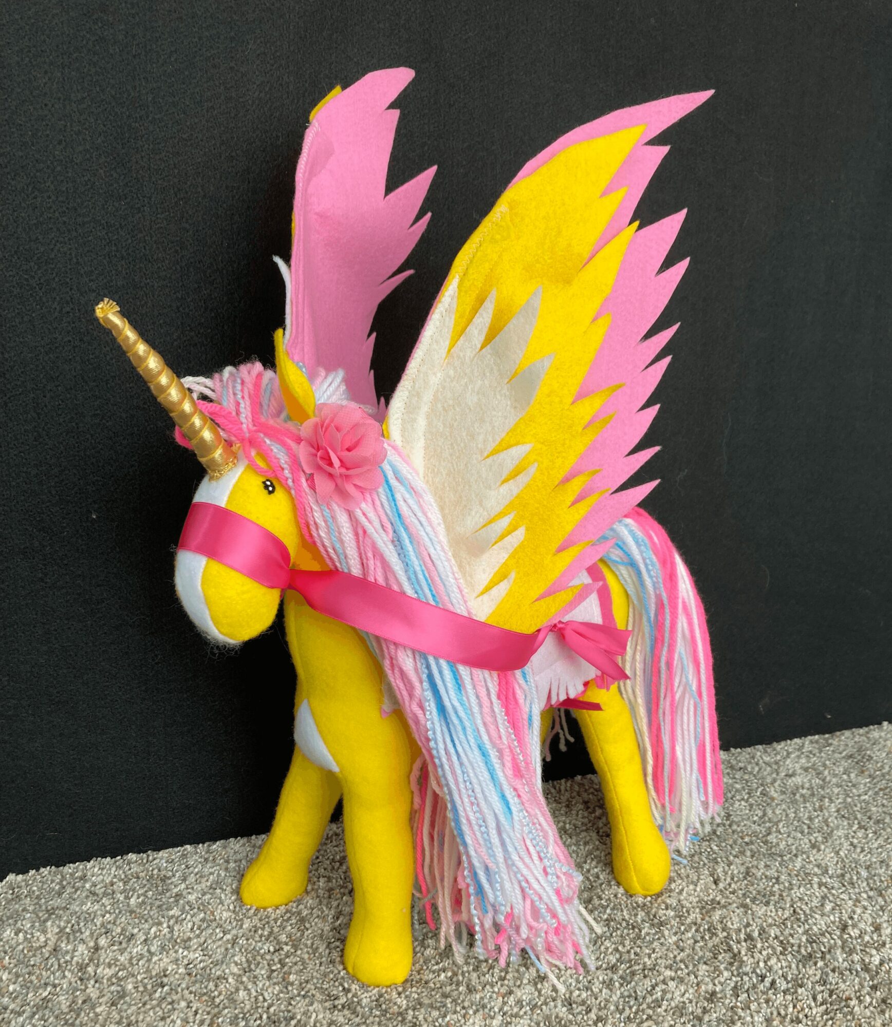 Colorful plush unicorn with wings, a horn, and a rainbow mane and tail, standing on a gray surface against a black background.