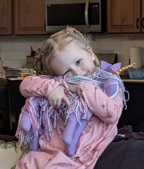 Young girl with braided hair hugs a purple stuffed animal, resting her head on it in a kitchen setting.