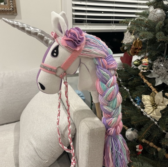 Colorful plush unicorn head with a rainbow mane and horn, hanging on a chair near a decorated Christmas tree.