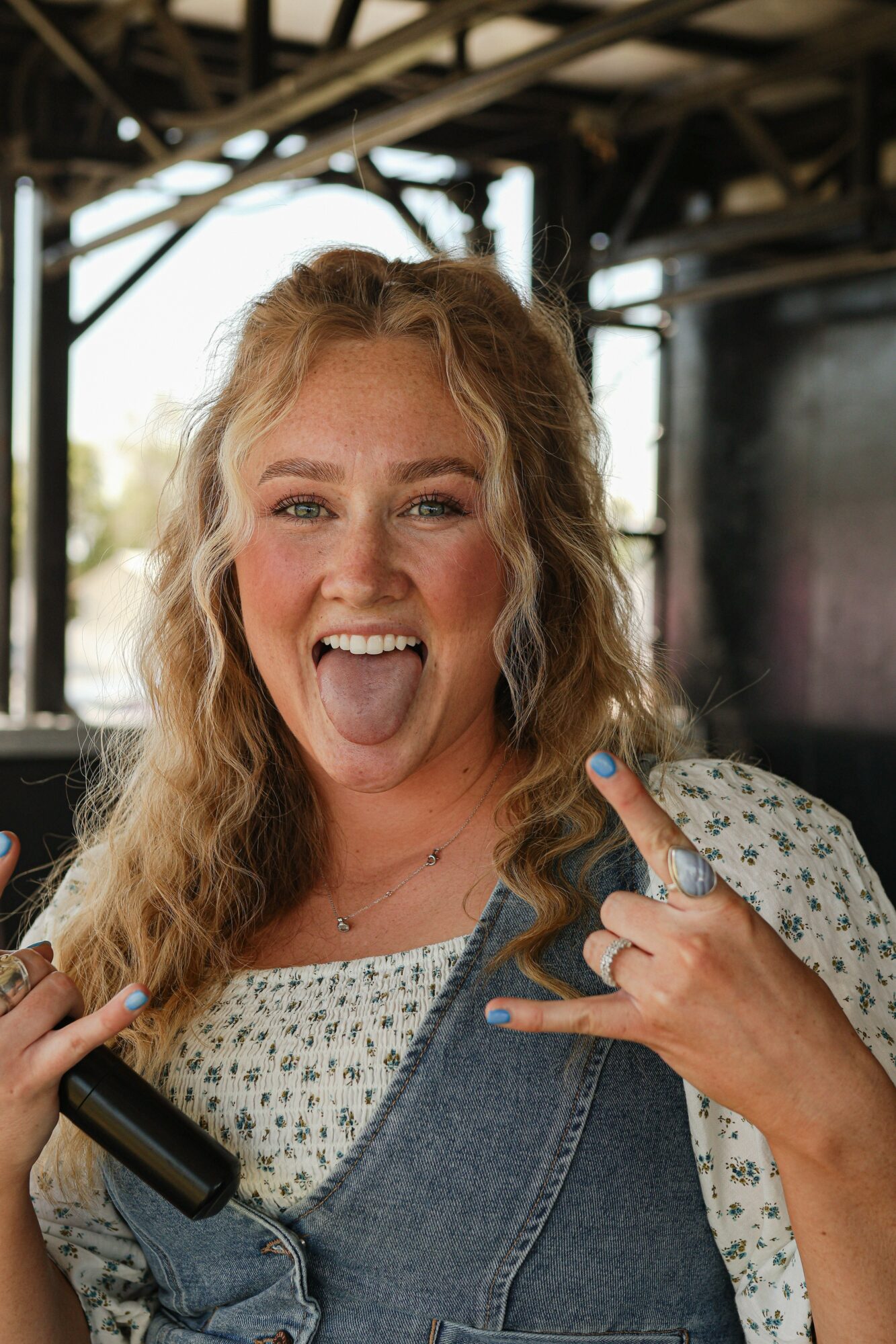 Young woman with curly hair sticking out her tongue, making rock hand gestures, wearing a polka dot shirt and denim overalls, smiling.