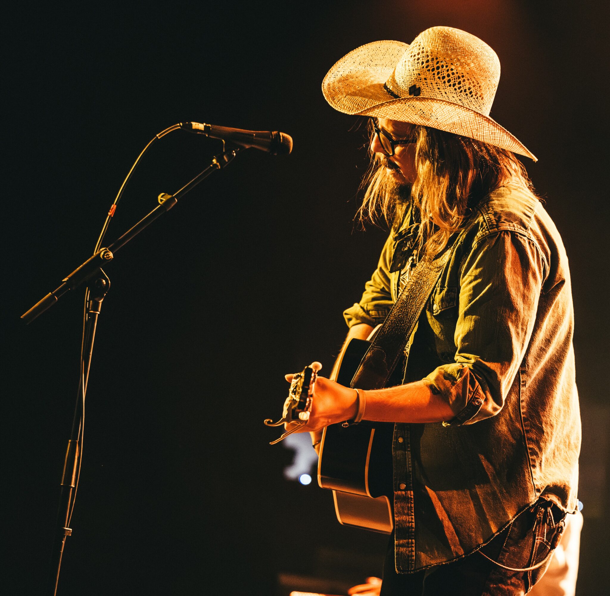 Person wearing a large hat playing guitar on stage with a microphone, illuminated by warm lighting.