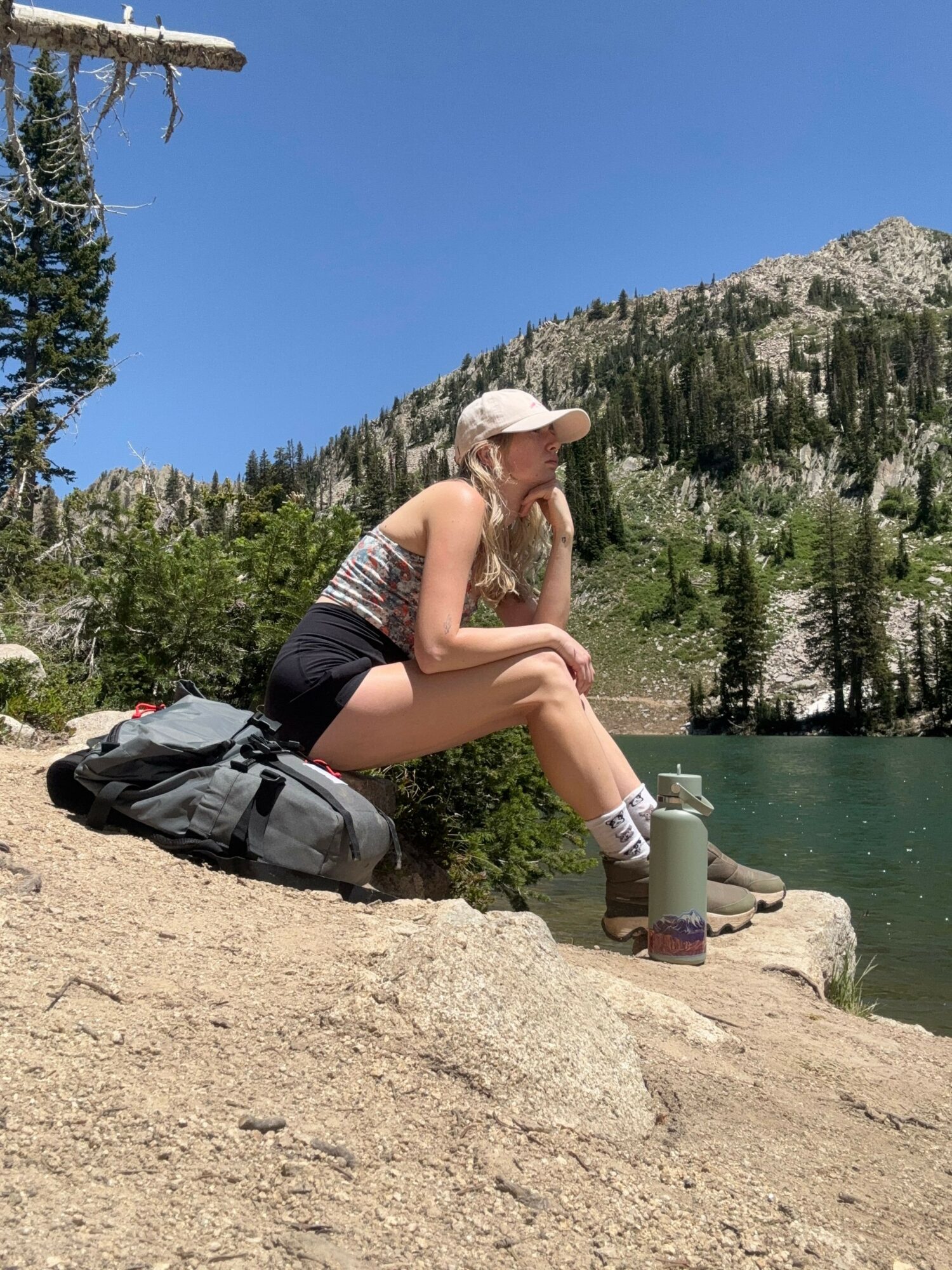 Person sitting on a rock near water, holding a drink, with a backpack beside them, trees and mountains in background.