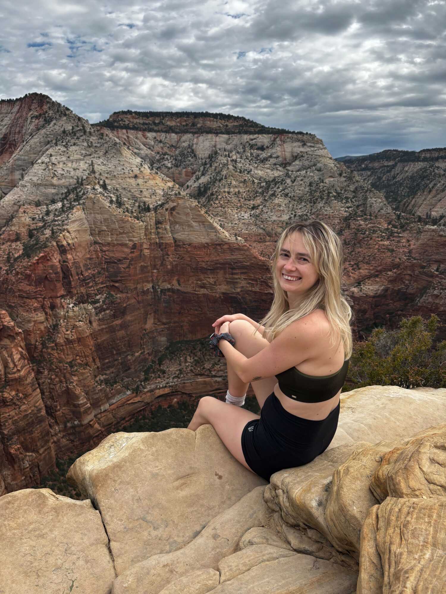 Woman sitting on rocks outdoors with a canyon and cloudy sky in background.