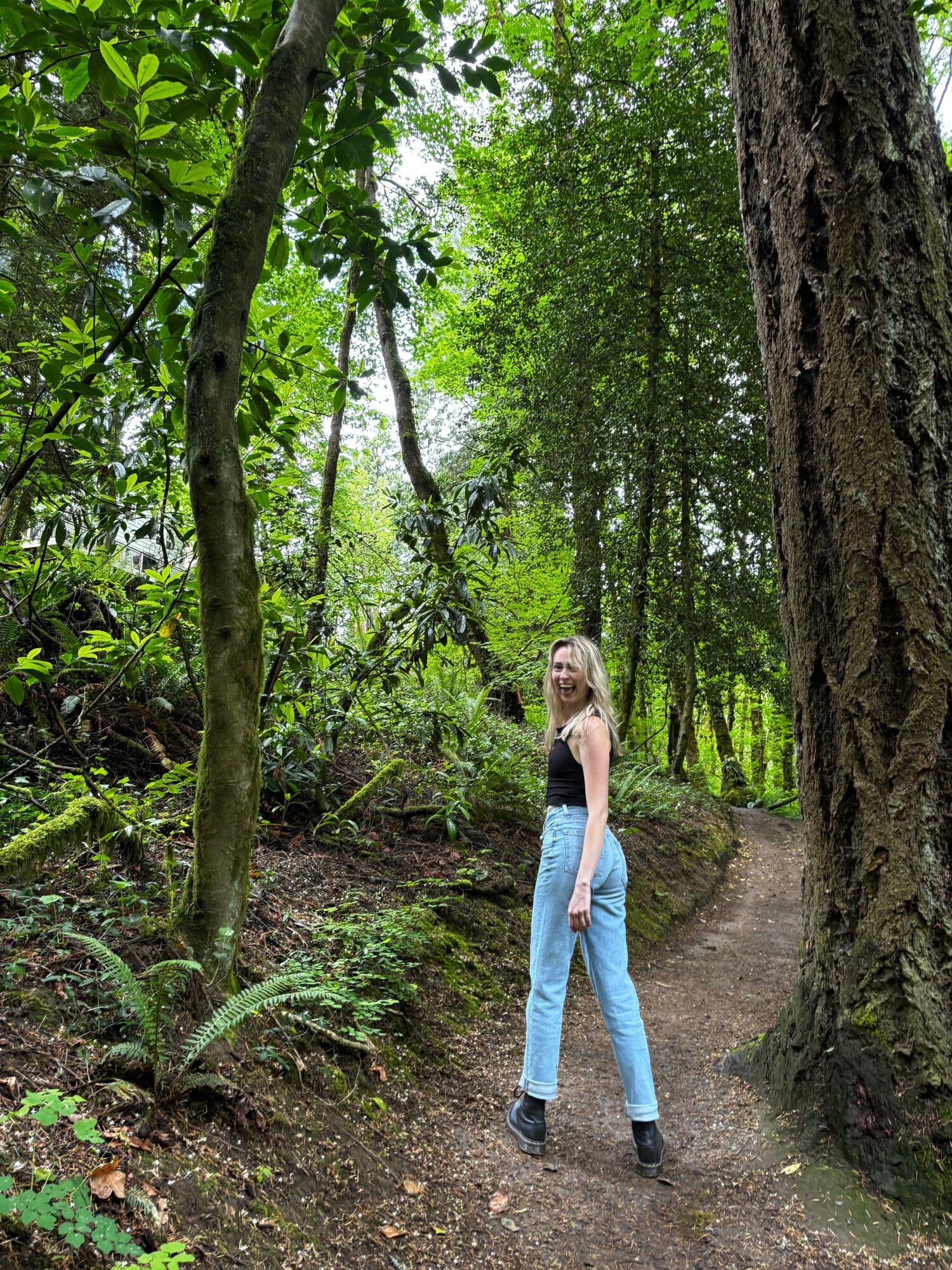 Woman standing on a forest trail surrounded by green trees and plants, with sunlight filtering through leaves.