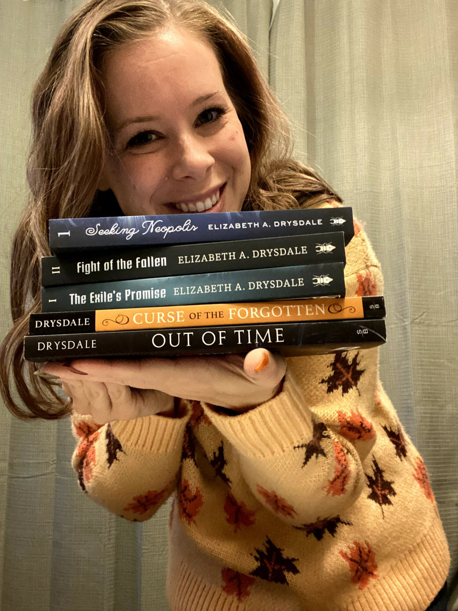 Person with long hair smiling, holding a stack of books, wearing a sweater with a leaf pattern, standing in front of curtains.