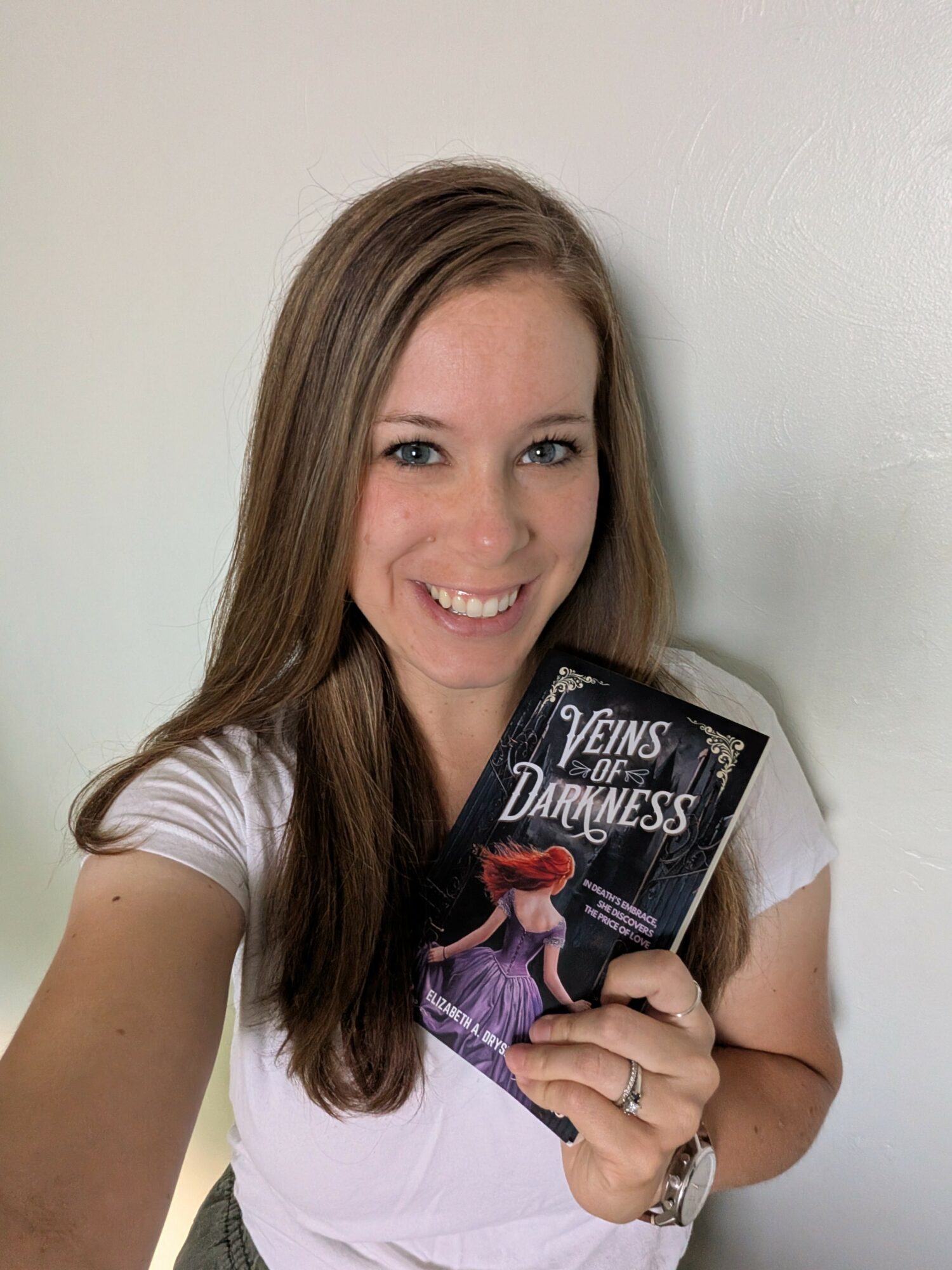 Smiling woman holding a book titled 'Veins of Darkness' in front of a plain wall.