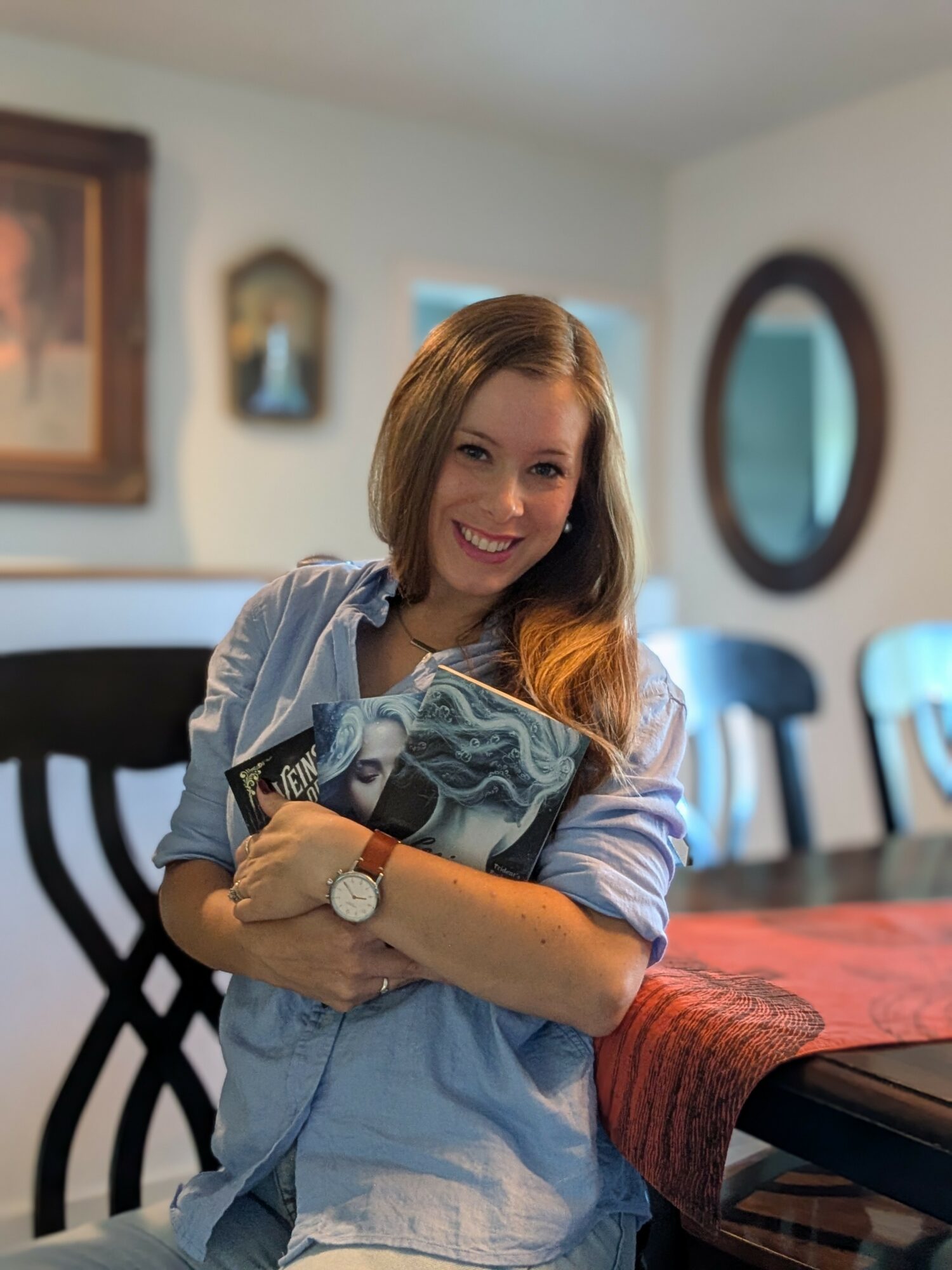 Young woman with shoulder-length hair smiling, holding a magazine, in a dining room with chairs and wall decorations