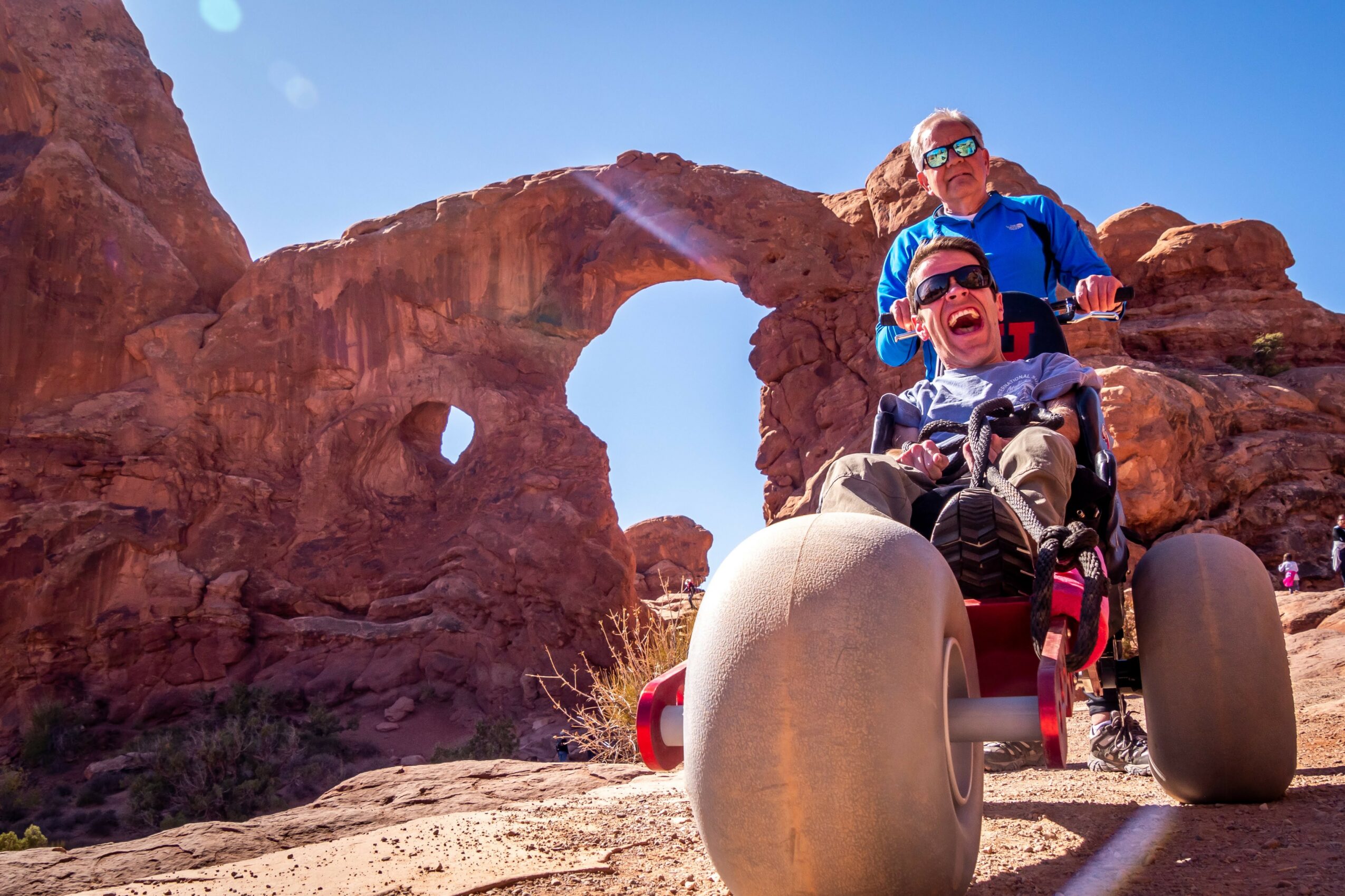 Two people in a wheelchair on a rocky landscape with natural arches in the background, smiling and enjoying the outdoors.