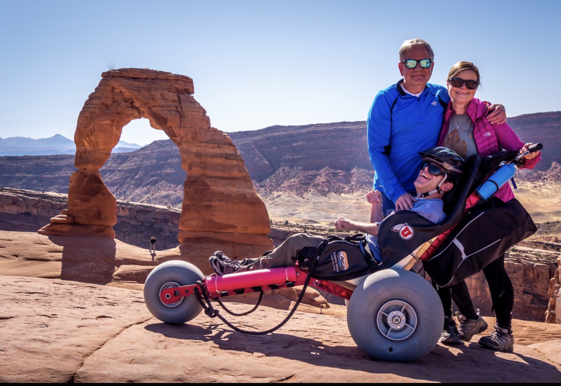 Three people with a wheelchair in a desert landscape with a natural rock arch in the background.