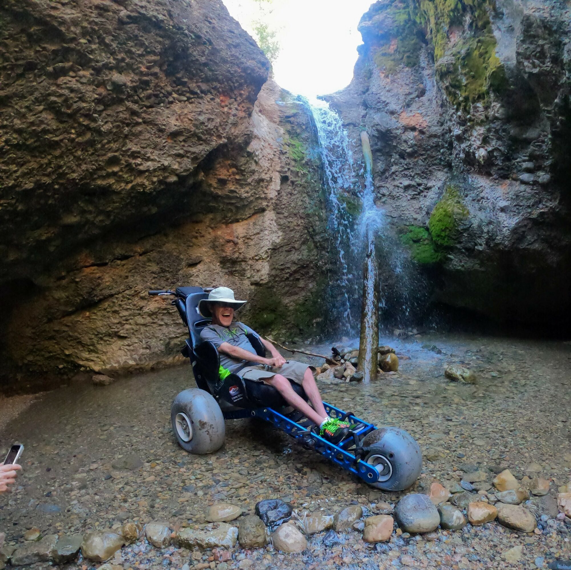 Child in a wheelchair on rocky ground near a waterfall in a canyon, wearing a hat and sunglasses.