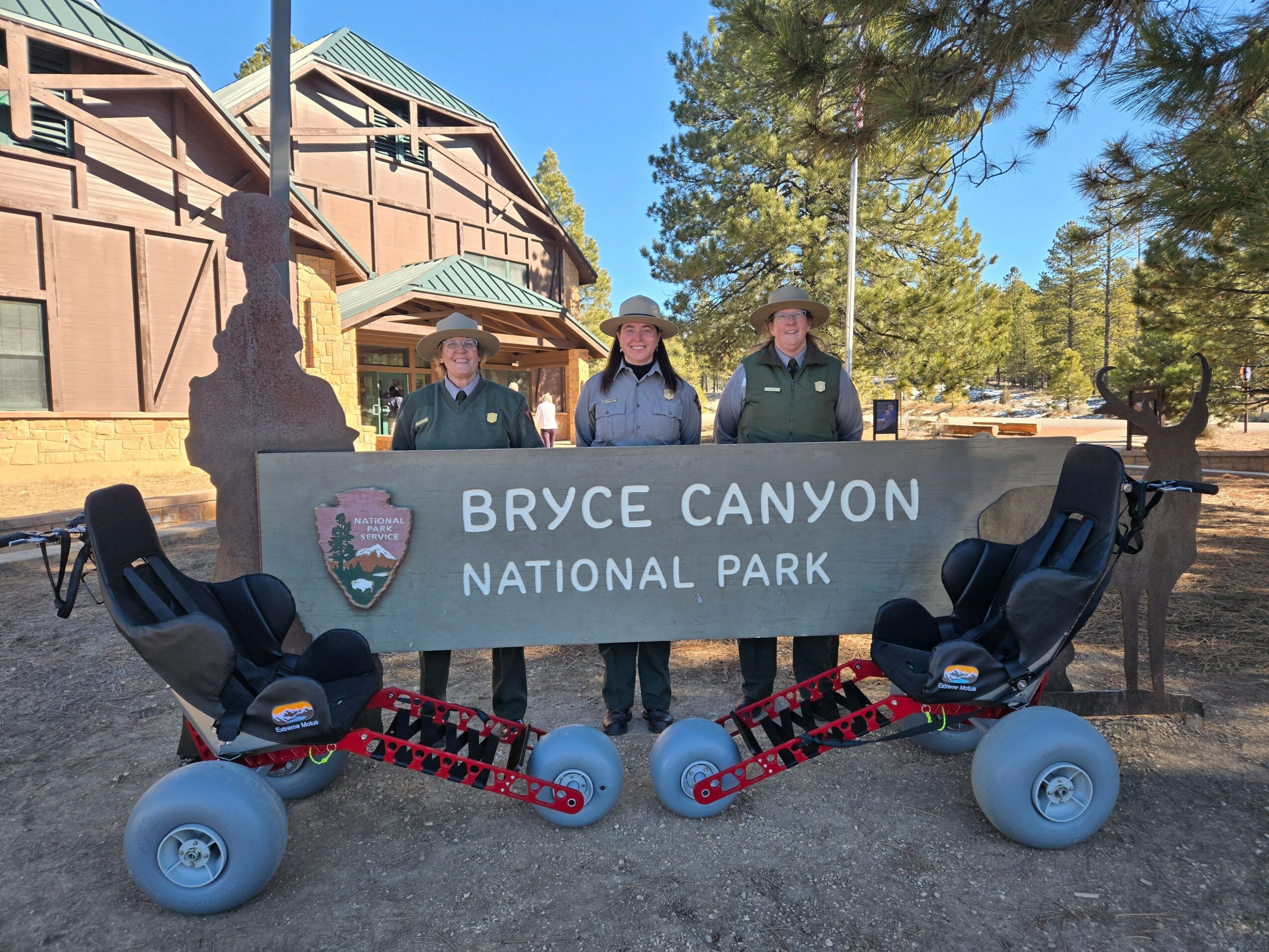 Three park rangers stand behind a large sign reading Bryce Canyon National Park, with two skateboards in front, trees and buildings in background.