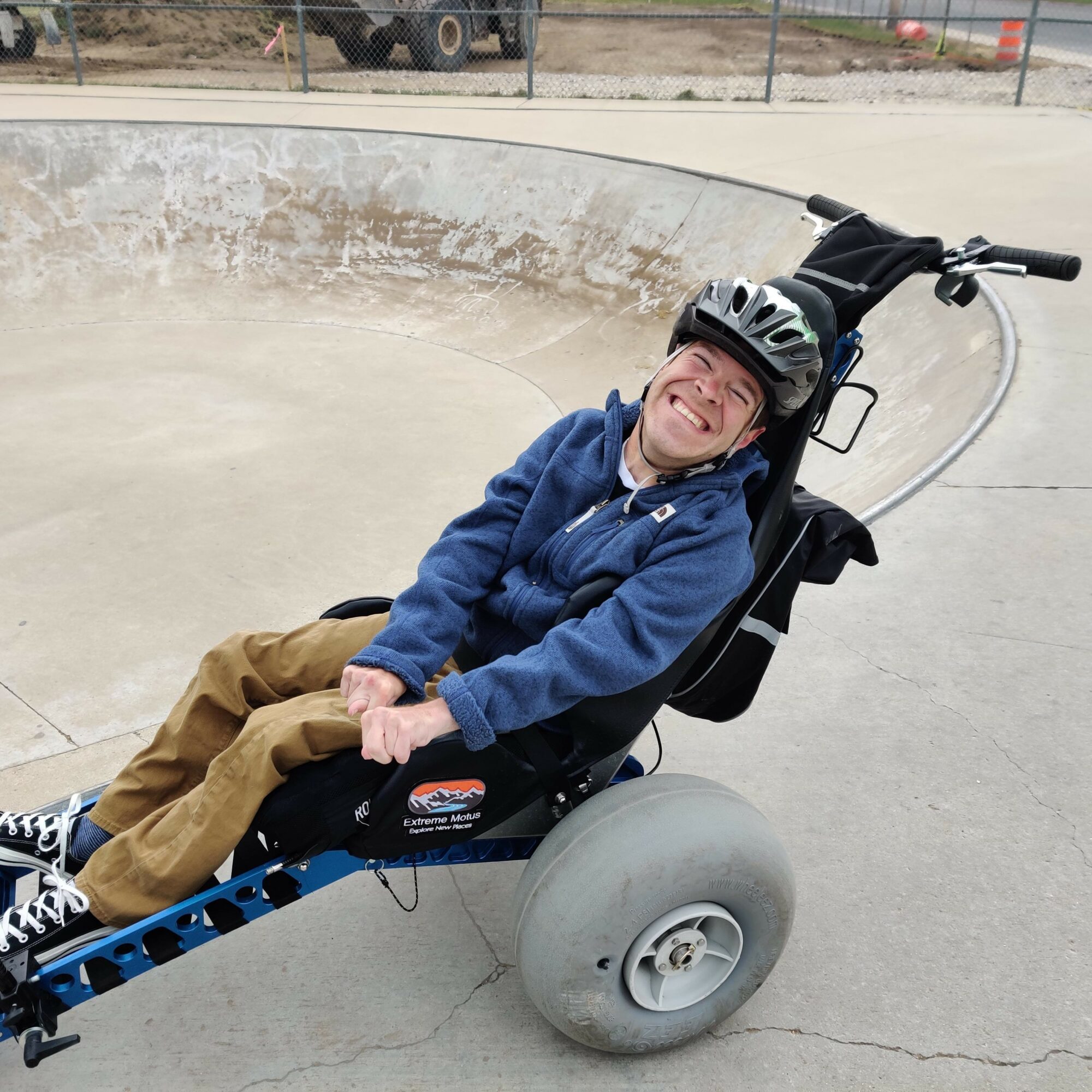 Person smiling and wearing a helmet, sitting in a wheelchair at a skate park bowl, holding a skateboard, with a fence and vehicles in background.
