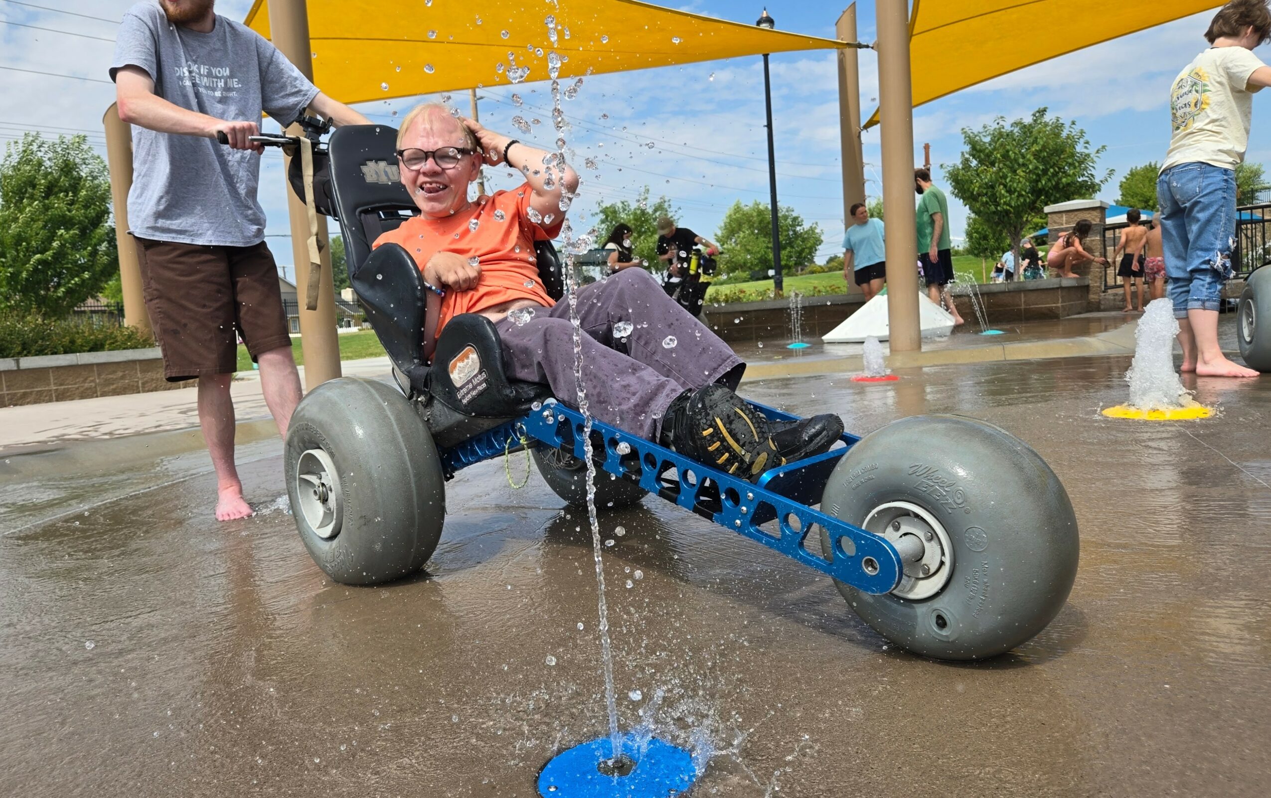 Child in a go-kart at a splash pad, water spraying from a fountain, other children playing nearby, sunny day.
