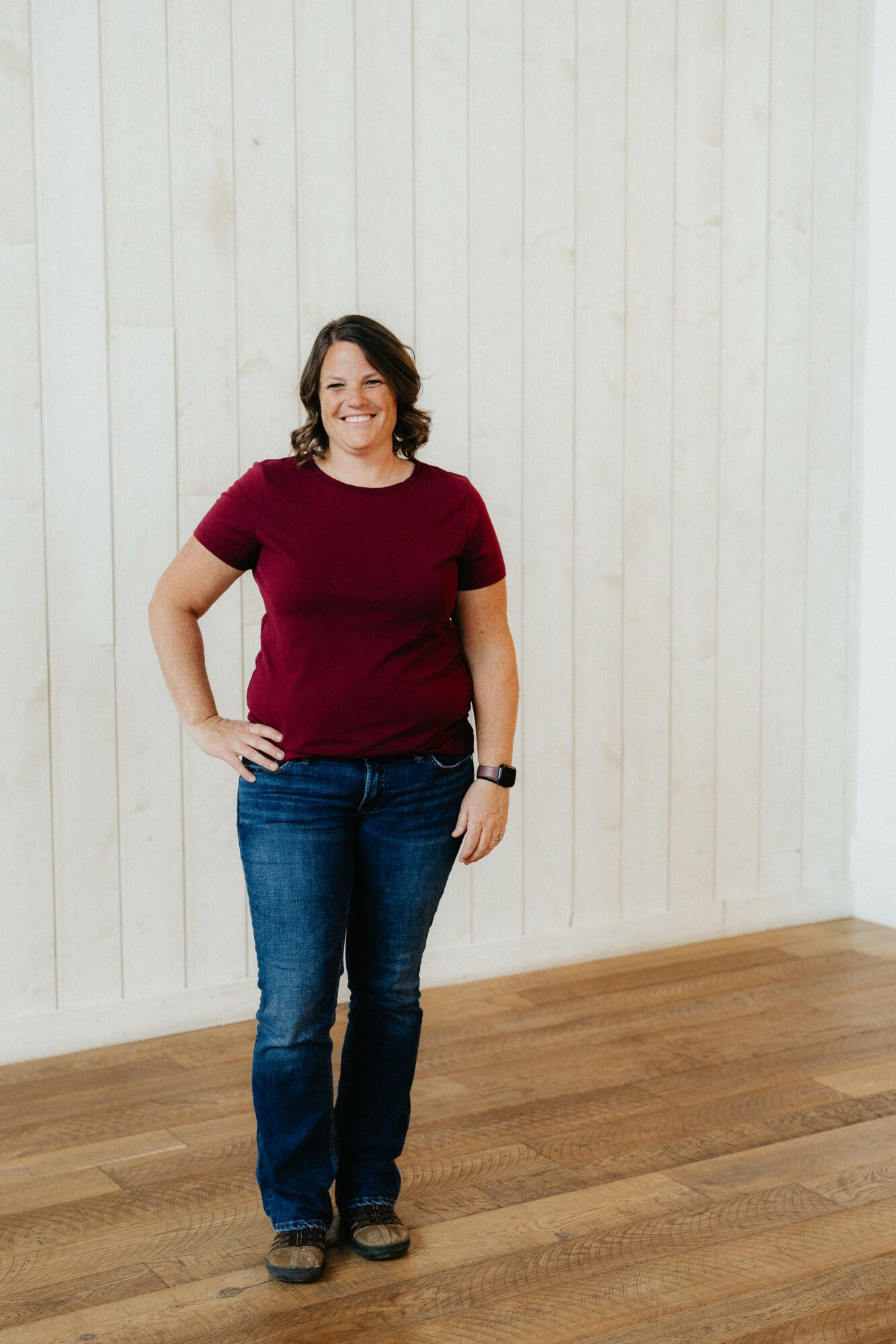 Woman standing indoors with a white wall background, smiling, wearing a red shirt and jeans.