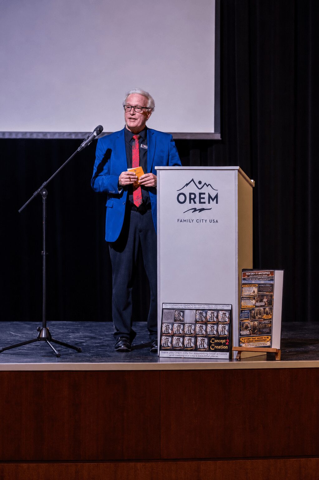 Older person with glasses in a blue blazer stands on stage behind a podium, holding a yellow object, with a microphone nearby.
