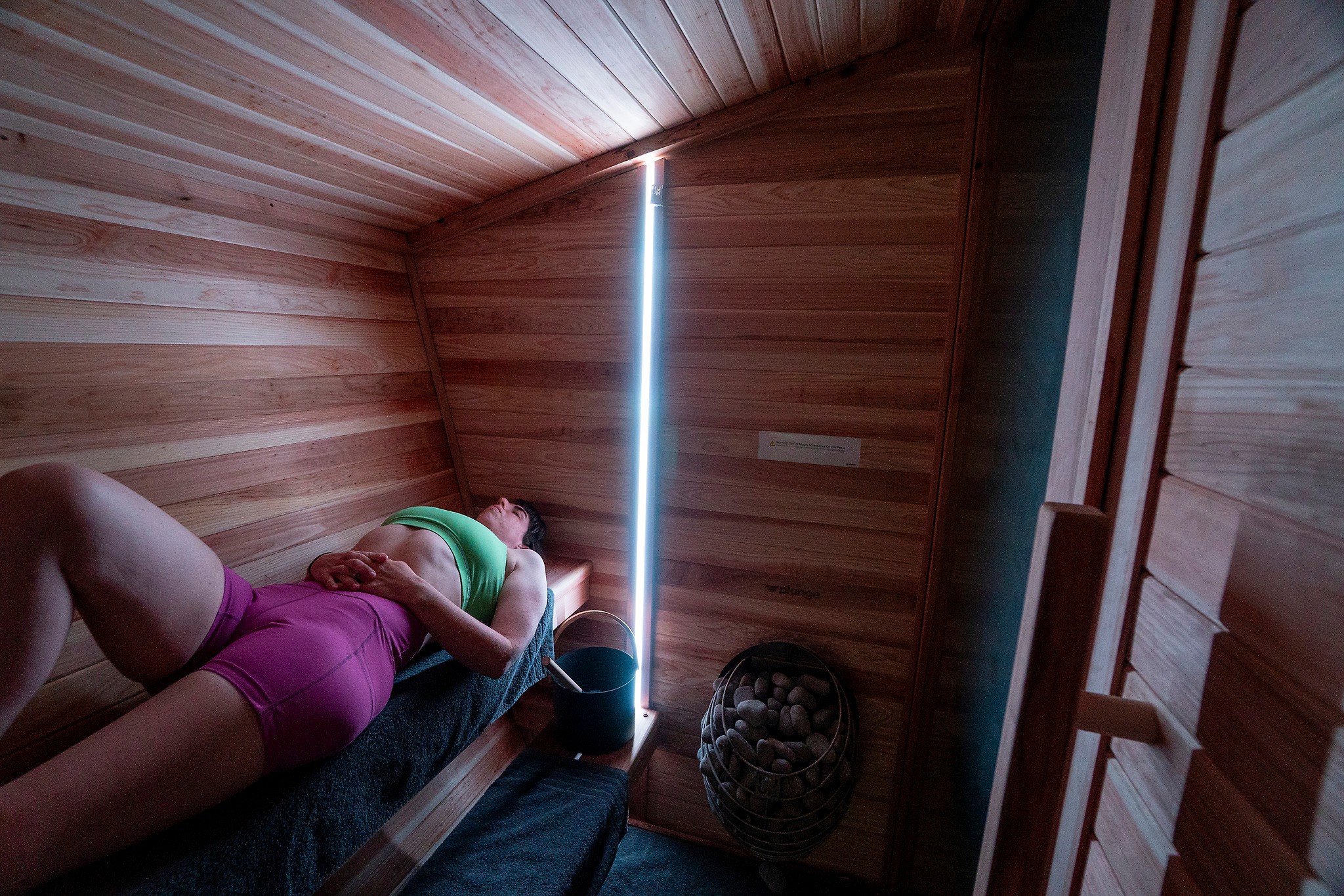 Person lying on bed in a wooden room, illuminated by a vertical light strip, with a decorative vase nearby.