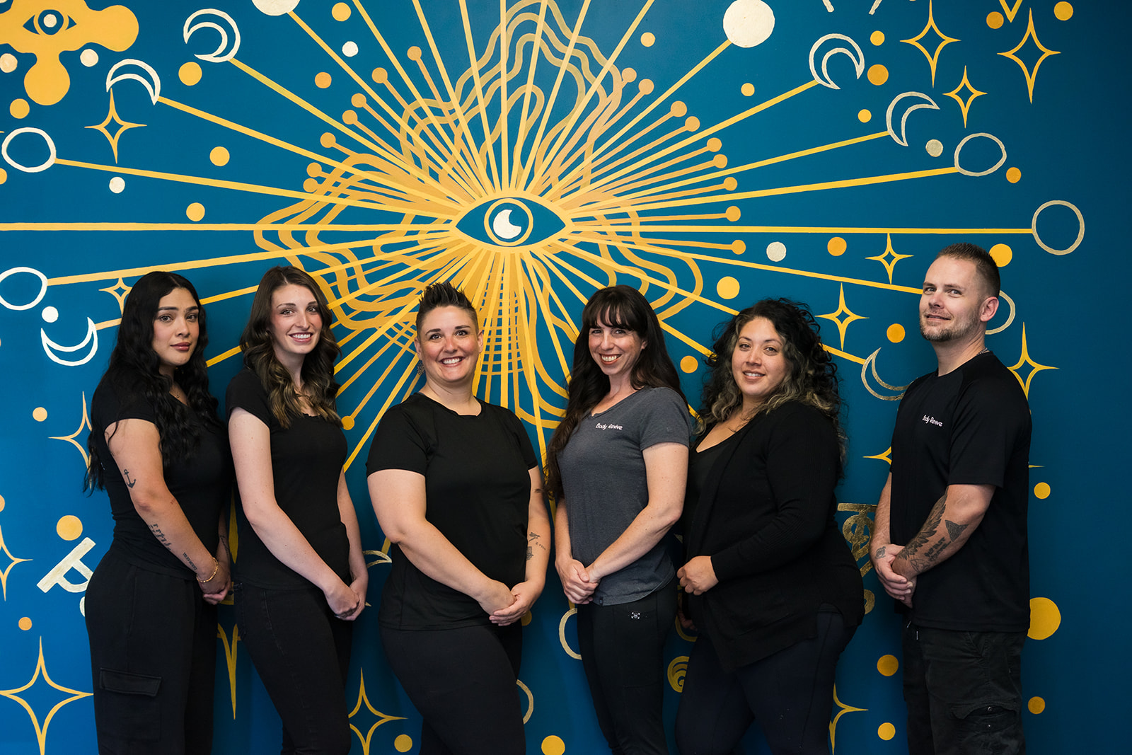 Six people stand in front of a blue background with a gold eye and radiating lines design.