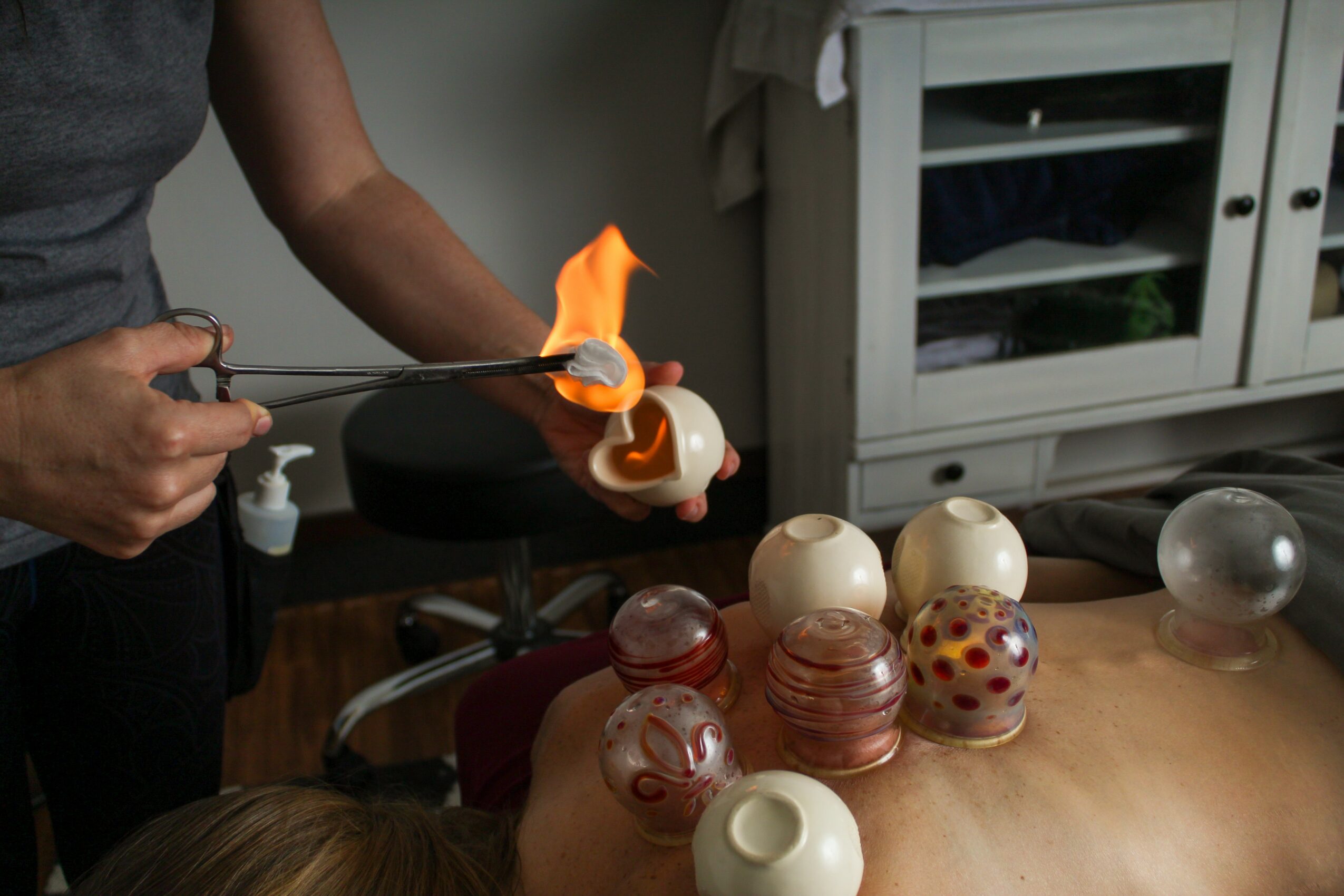 Person torching glass objects with a flame, various glass art pieces on a table, cabinet in background.