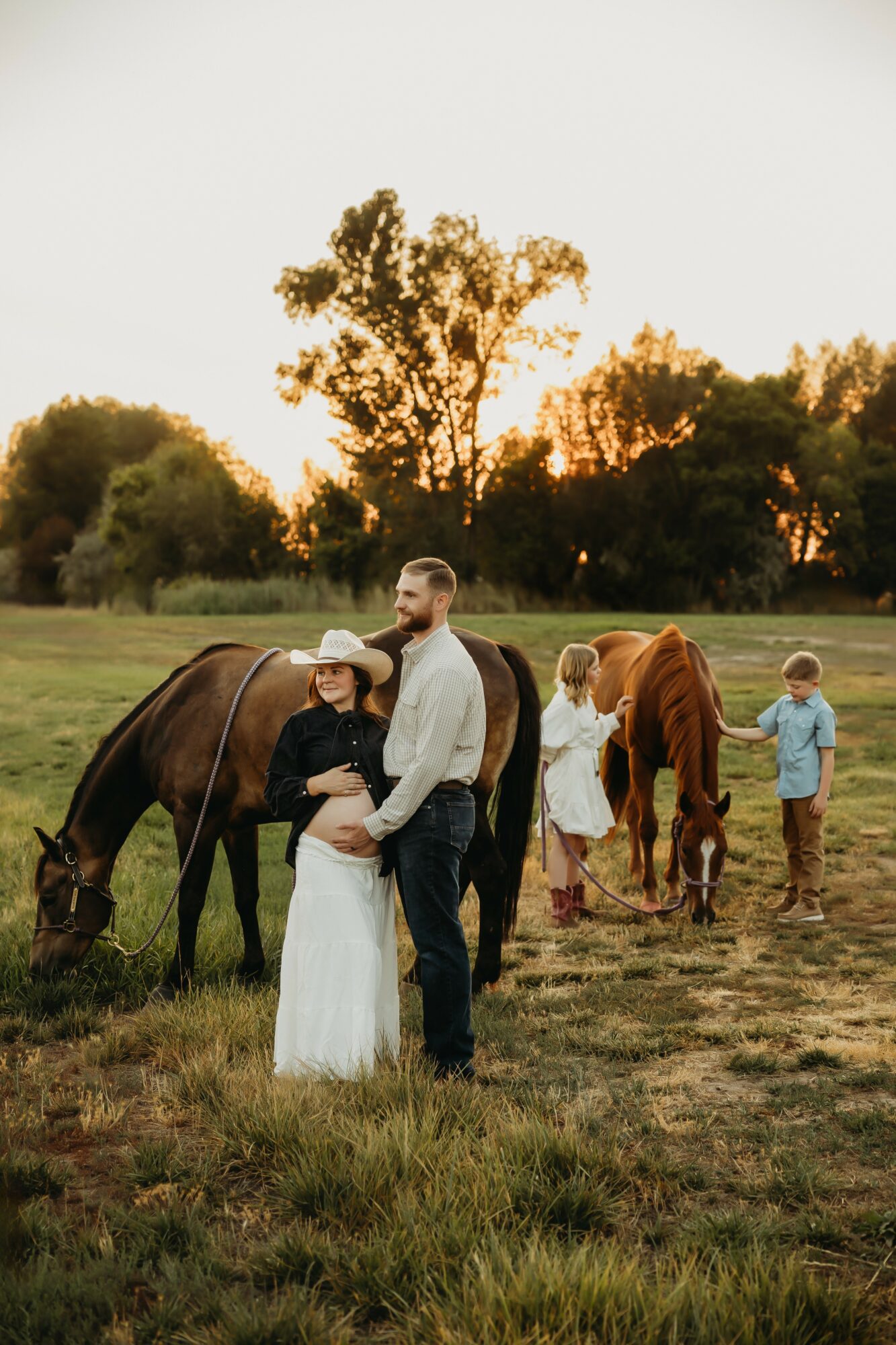 Couple standing in a field with horses, children nearby, trees and sunset in background.