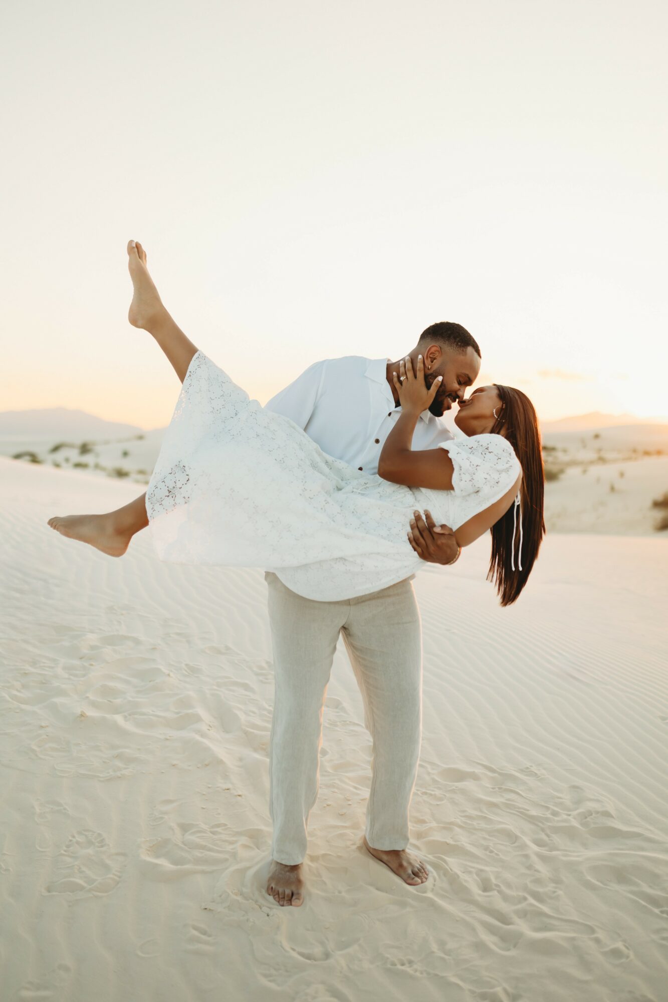 A couple in white clothing dancing on sand dunes, the man lifting the woman, both smiling, with a clear sky background.