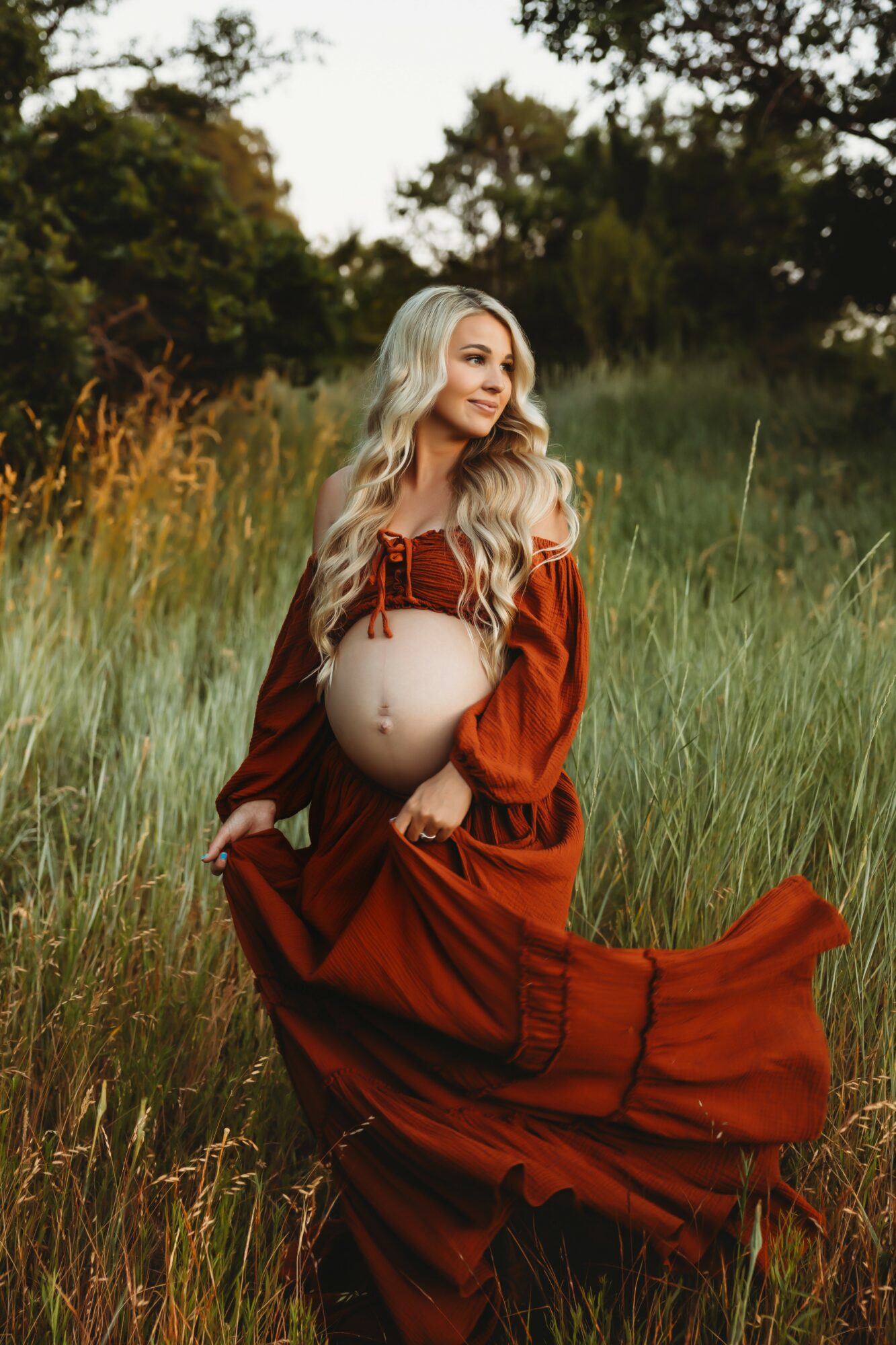 Pregnant woman in a red dress sitting in a grassy field with trees in the background, holding her belly.