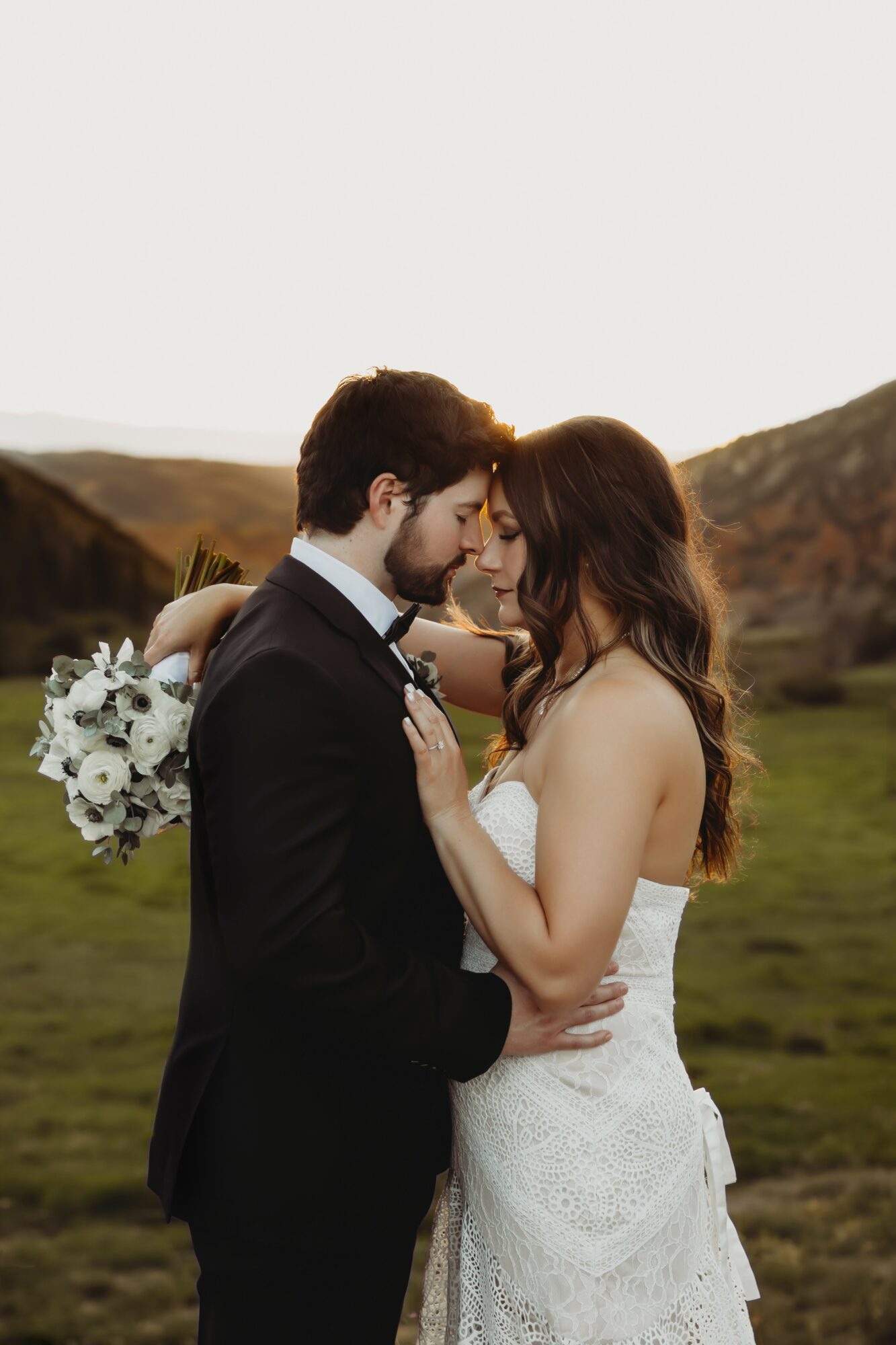 A couple in wedding attire standing close outdoors, with a landscape of hills in the background.