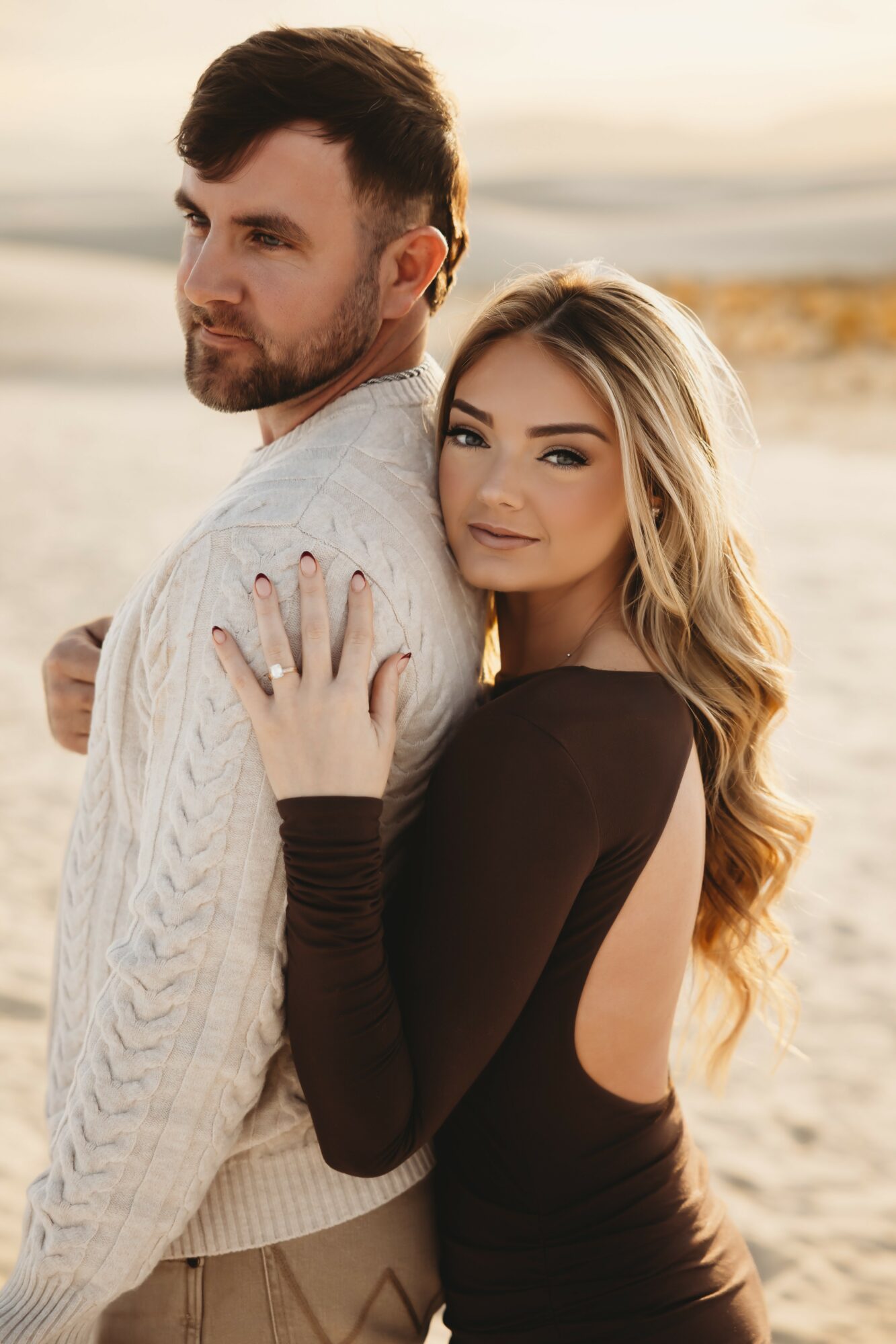 A couple standing on a beach at sunset, with the woman smiling and embracing the man from behind.