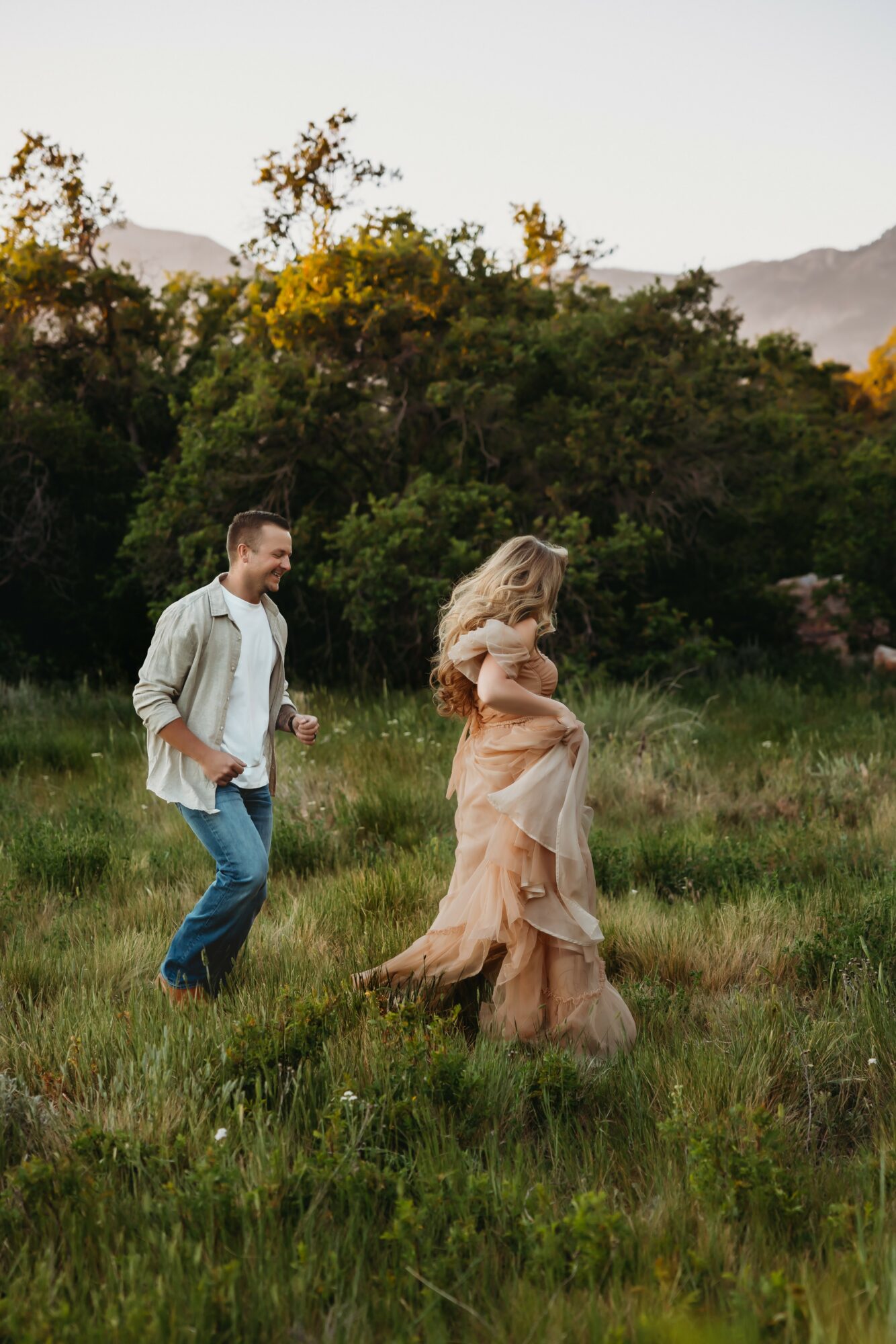 A man and woman walk through a grassy field with trees and mountains in the background, woman in a flowing dress.