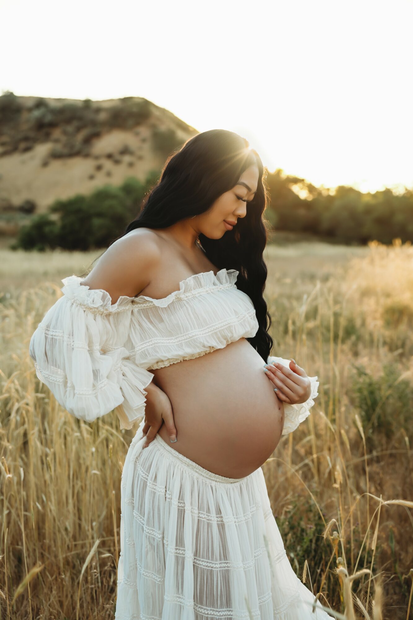 Pregnant woman in white dress standing in a field with a hill in the background, sunlight shining behind her.