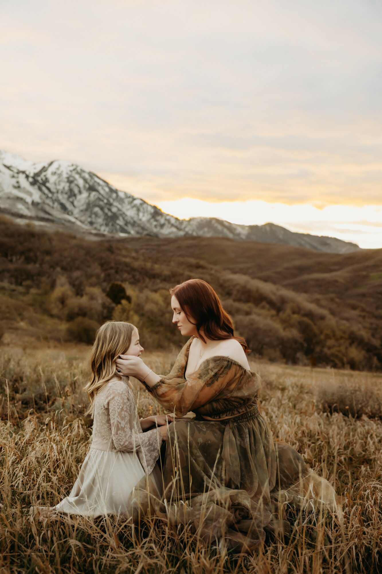 Woman and girl in a grassy field with mountains and snow in background, woman touching girl's face, sunset sky.