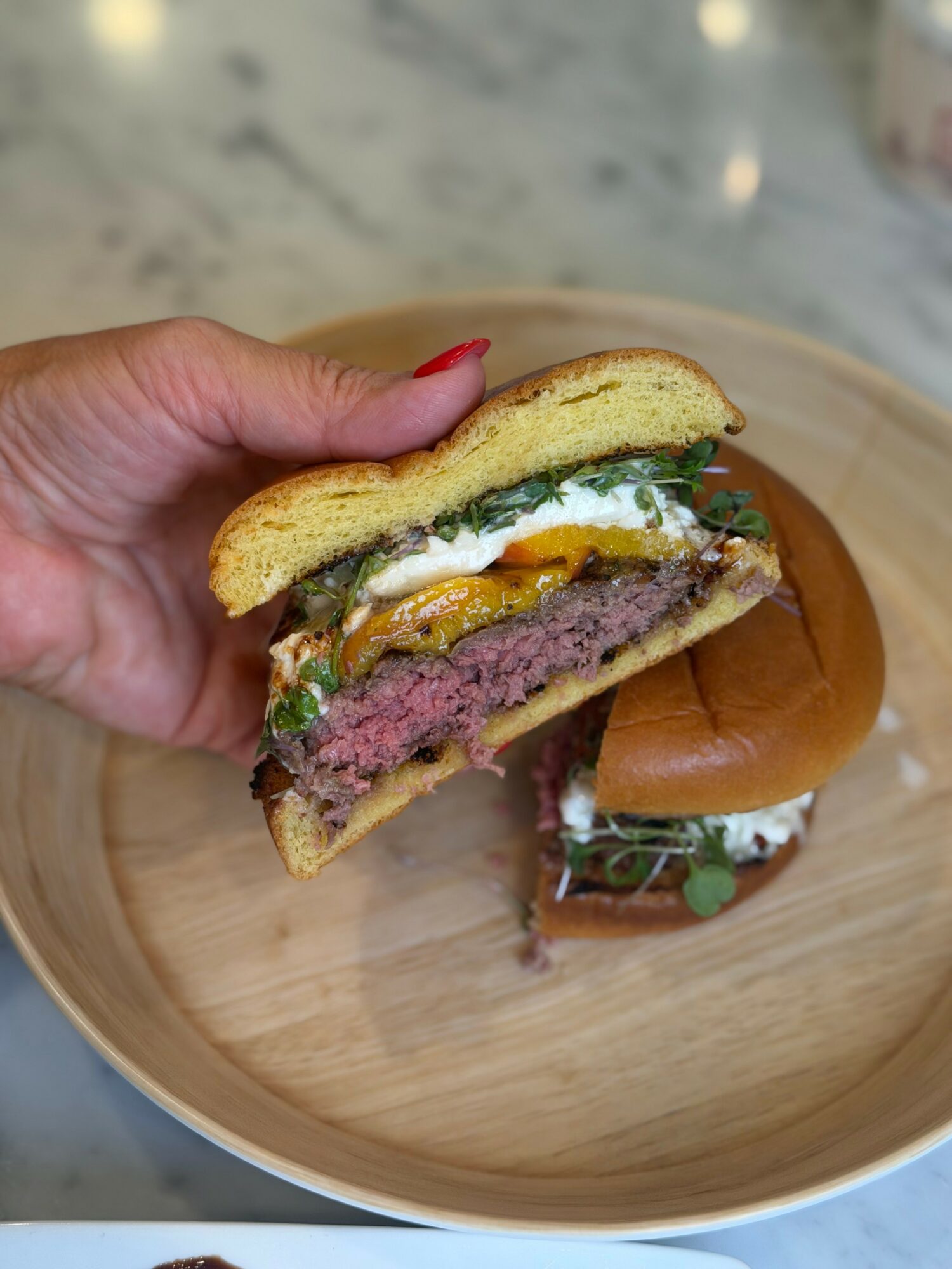 Hand holding a half-eaten burger with beef patty, cheese, lettuce, tomato, and sauce, on a wooden plate.