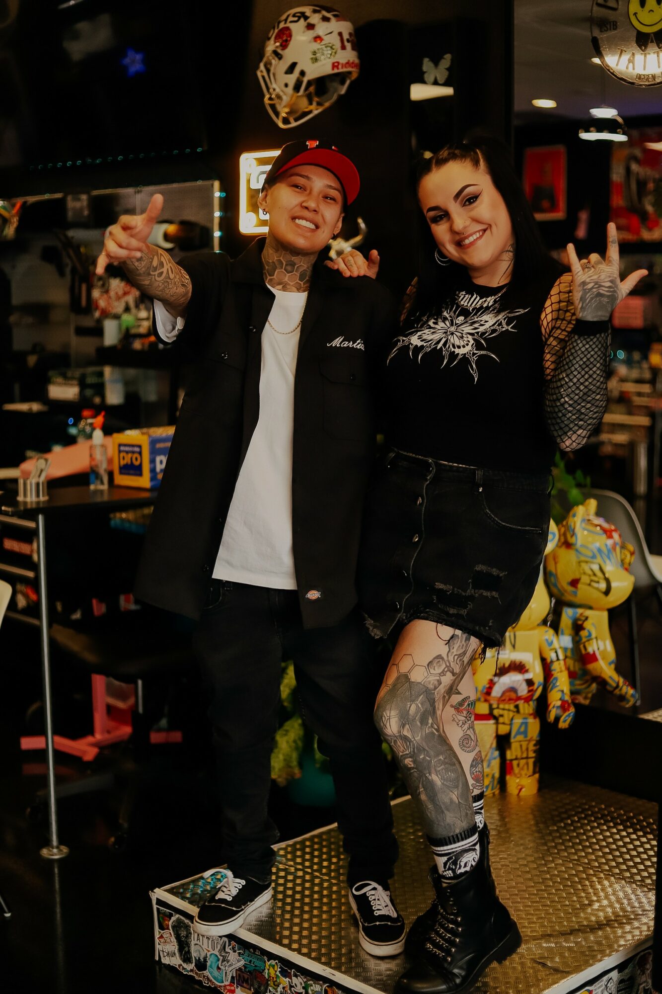 Two people smiling and posing in a store, surrounded by toys and merchandise, with one person wearing a red cap and tattoos.