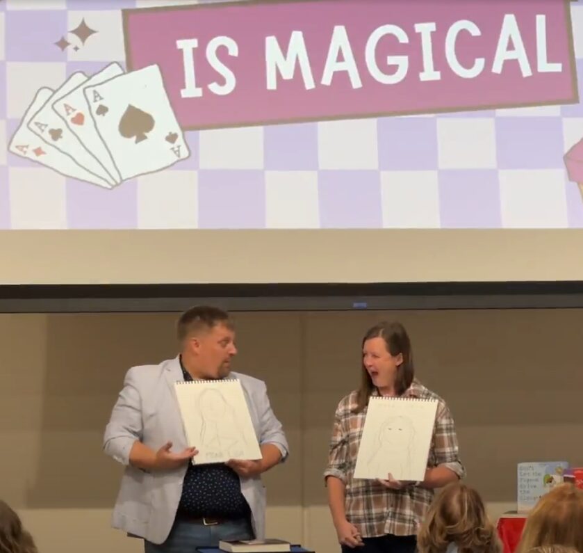 Two people standing on stage holding whiteboards, with a large screen behind them displaying a sign that says 'IS MAGICAL' and playing cards. Audience members are visible in the foreground.