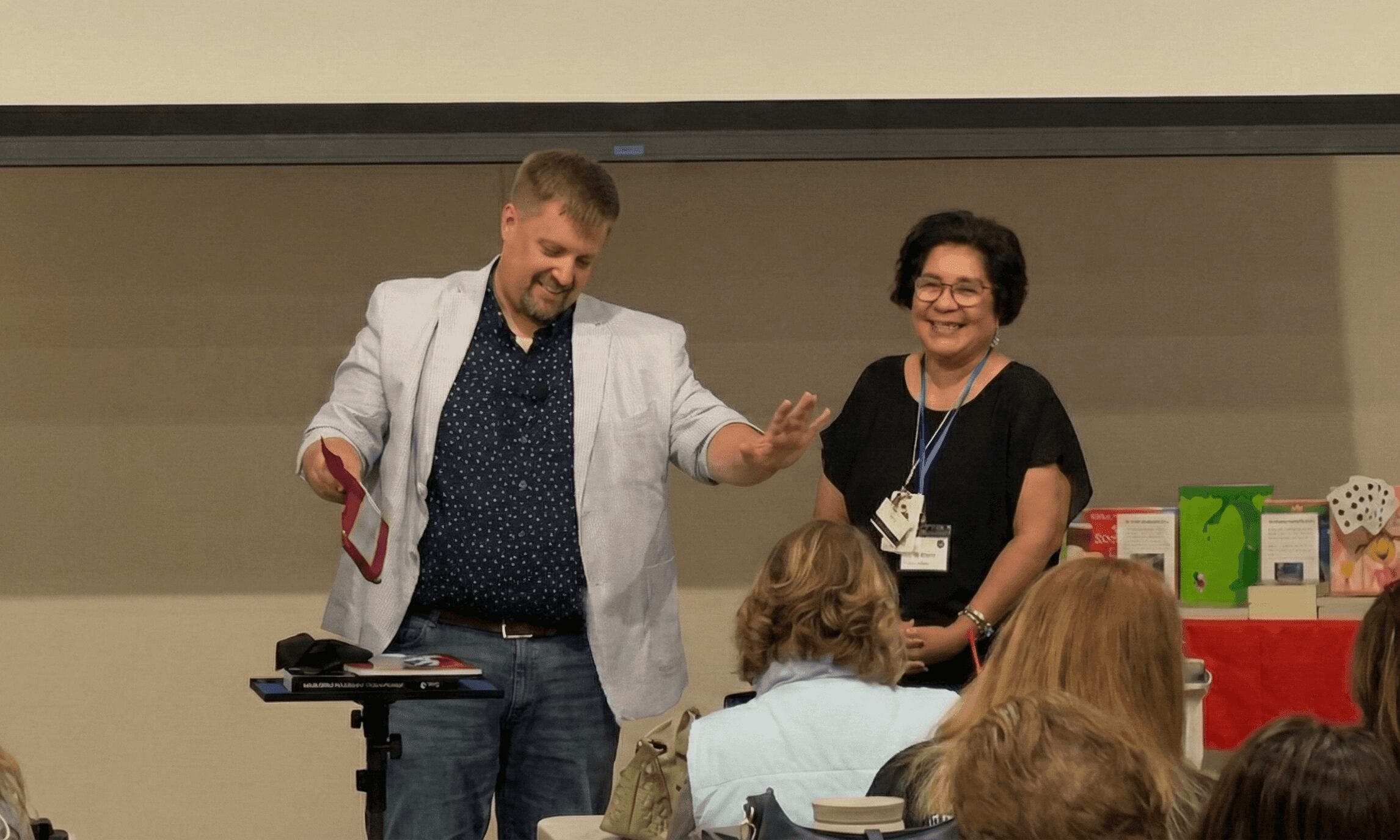 Man and woman standing on stage, smiling, with audience in foreground, in a conference room setting.