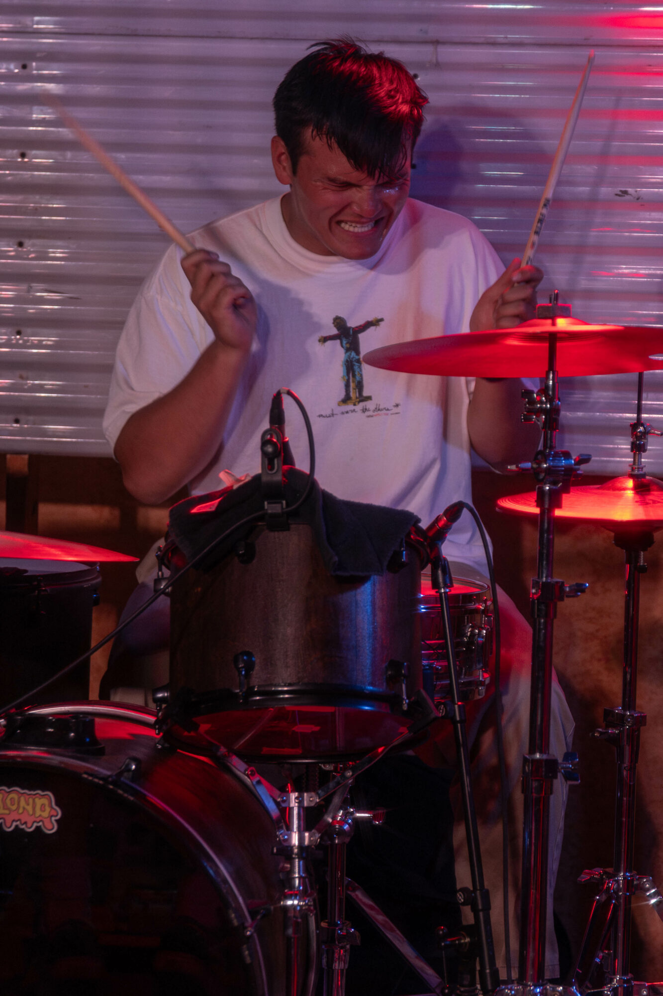 Person playing drums, smiling, holding drumsticks, with a drum set and red lighting.